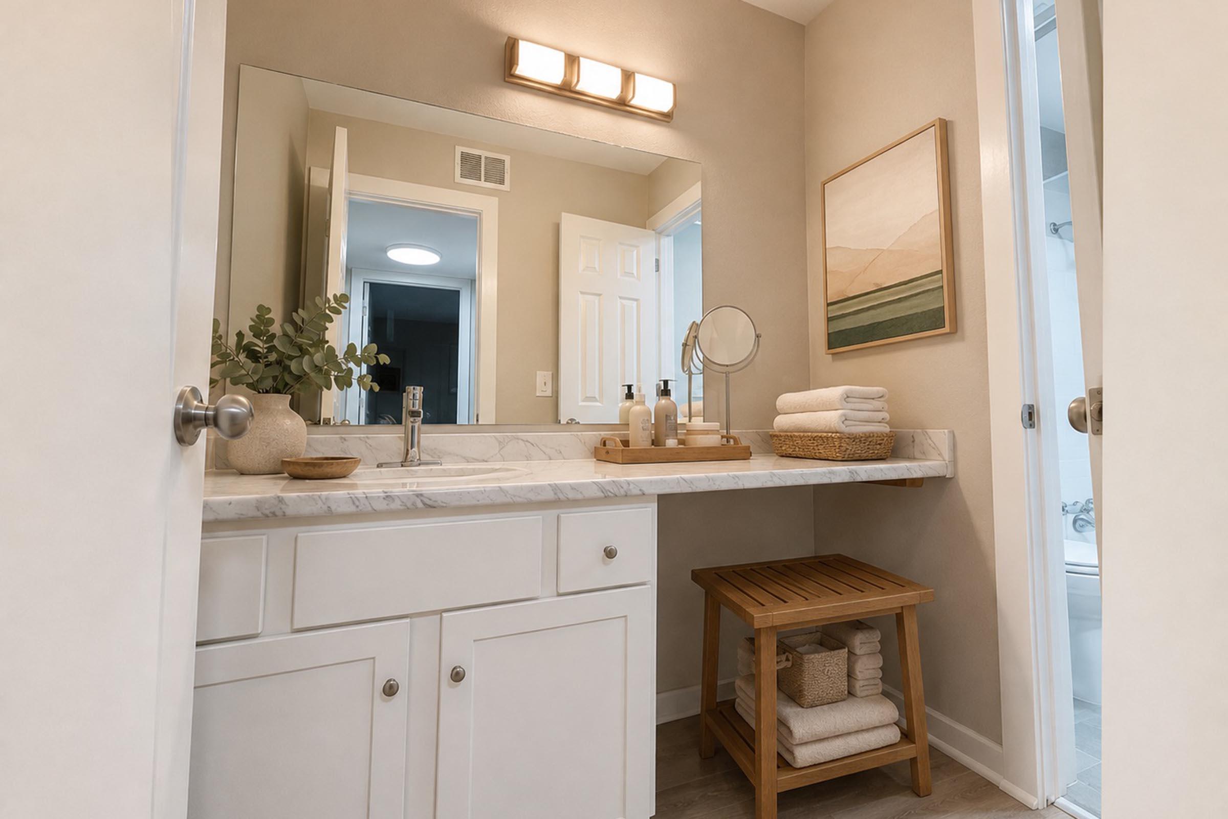 A modern bathroom featuring a beige color scheme. The countertop has neatly arranged toiletries, a round mirror, and a decorative plant. A wooden bench holds rolled towels, while a bright light fixture illuminates the space. A large mirror reflects the room, adding depth.