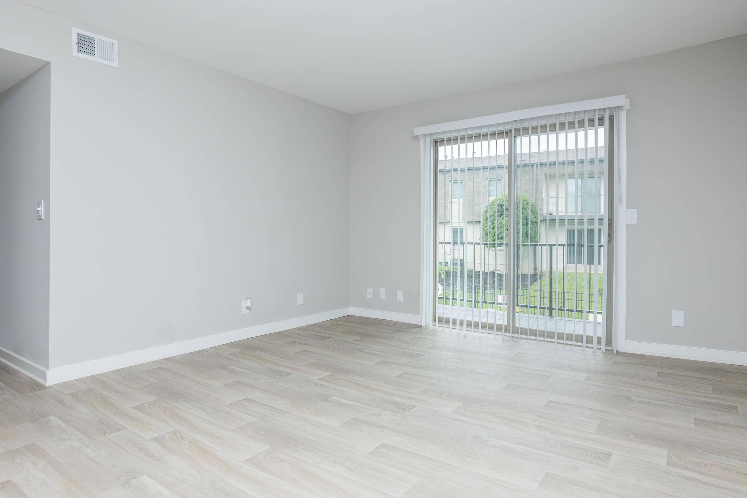 Spacious, empty room with light wood flooring and neutral gray walls. Large sliding glass door with white blinds leads to an outdoor area. Windows provide natural light, creating a bright and airy atmosphere. Ideal for a living space or office setup.