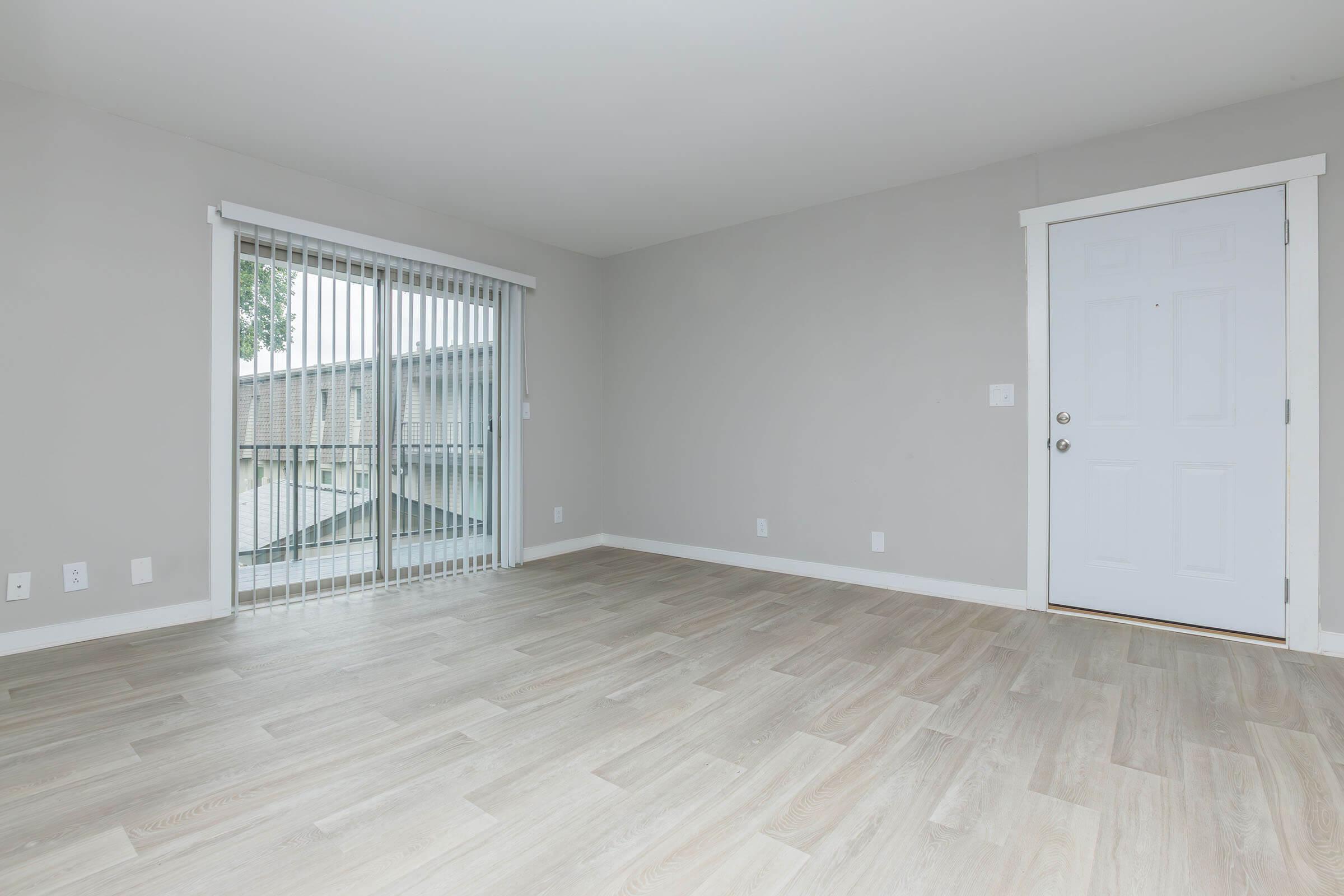 Spacious interior of a light-colored room featuring a large window with vertical blinds, revealing a view outside. The floor is composed of light wood-like planks, and there is a white front door on the right. The walls are painted in a neutral gray tone, creating a bright and airy atmosphere.