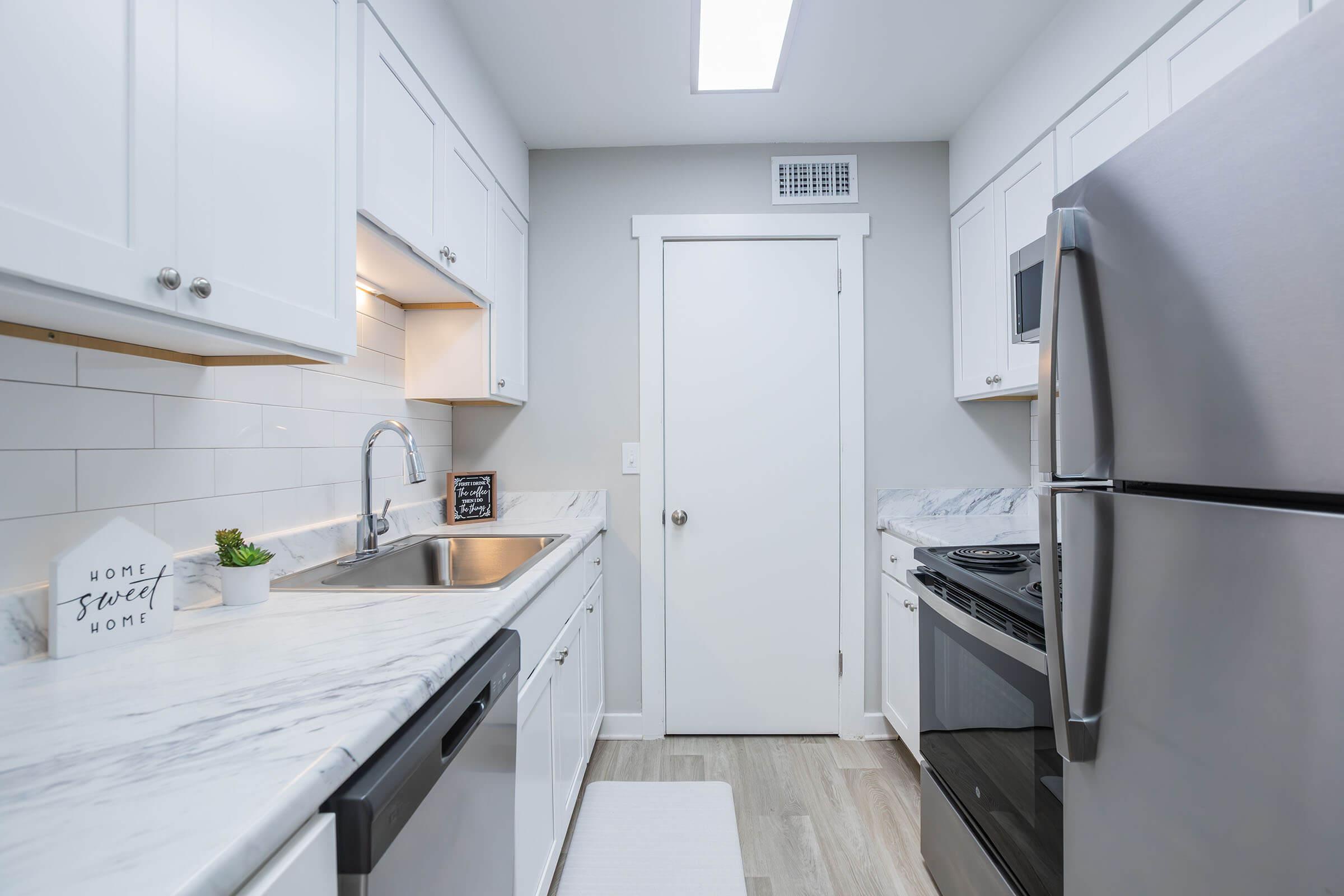 Modern kitchen with white cabinetry, stainless steel appliances, and marble countertops. The space features a sink, a stove, and a refrigerator. Natural light enters through a skylight, and a small decorative sign reading "Home Sweet Home" is visible on the countertop. The floor is light-colored wood.