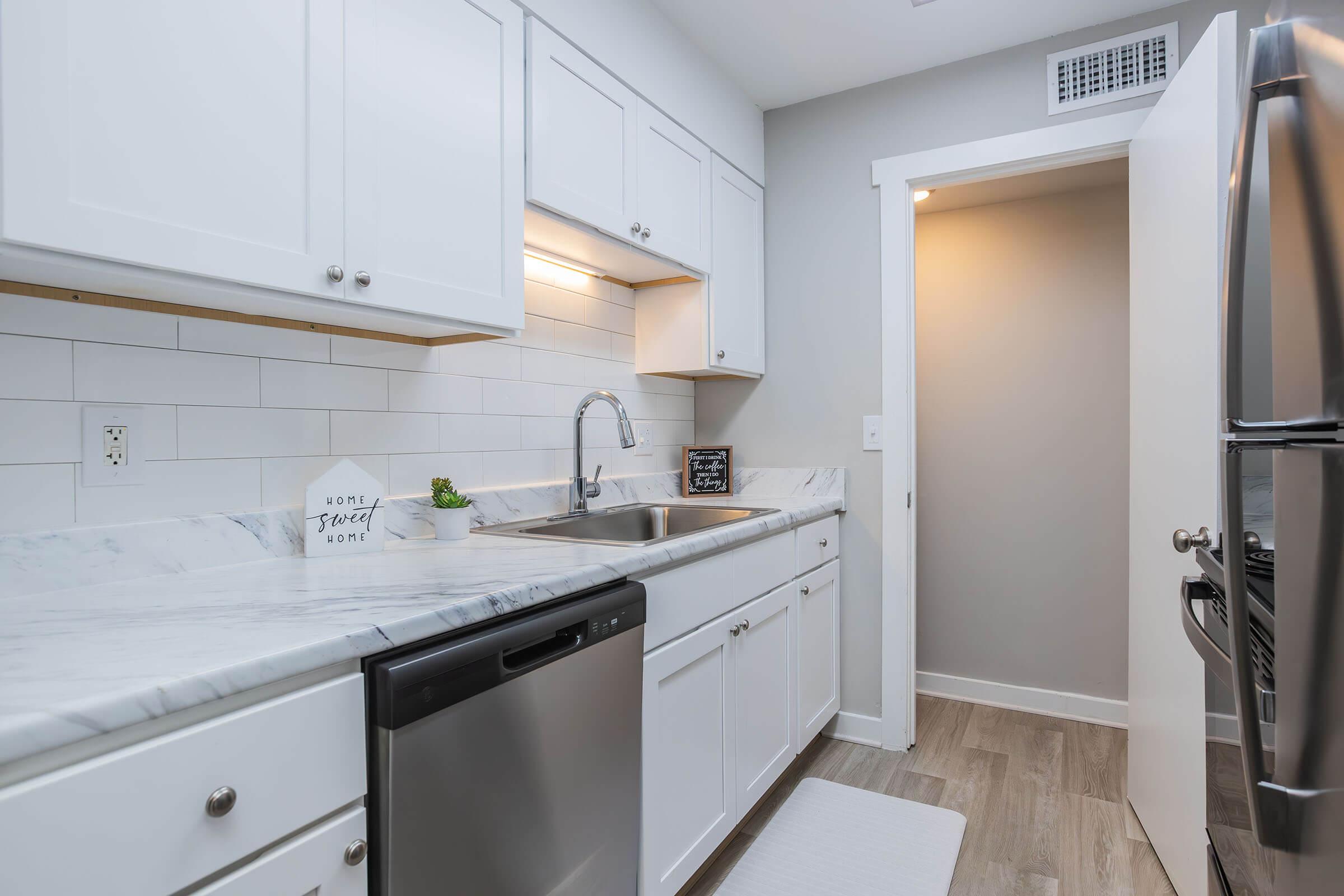 Modern kitchen with white cabinets, a marble countertop, and a stainless steel sink. Features include a dishwasher, a window, and light-colored walls. A door leads to another room, and there's a small decorative sign on the countertop. The floor has a light wood finish, adding a bright and clean ambiance.