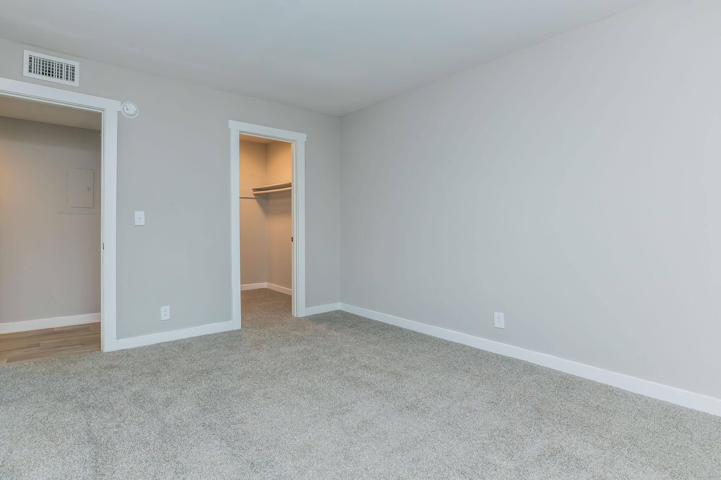 A spacious room with light gray walls and a plush carpet. There are two doors: one leading to a closet and another that opens into a hallway. The room is well-lit and offers a clean and simple design, suitable for various uses such as a bedroom or a home office.