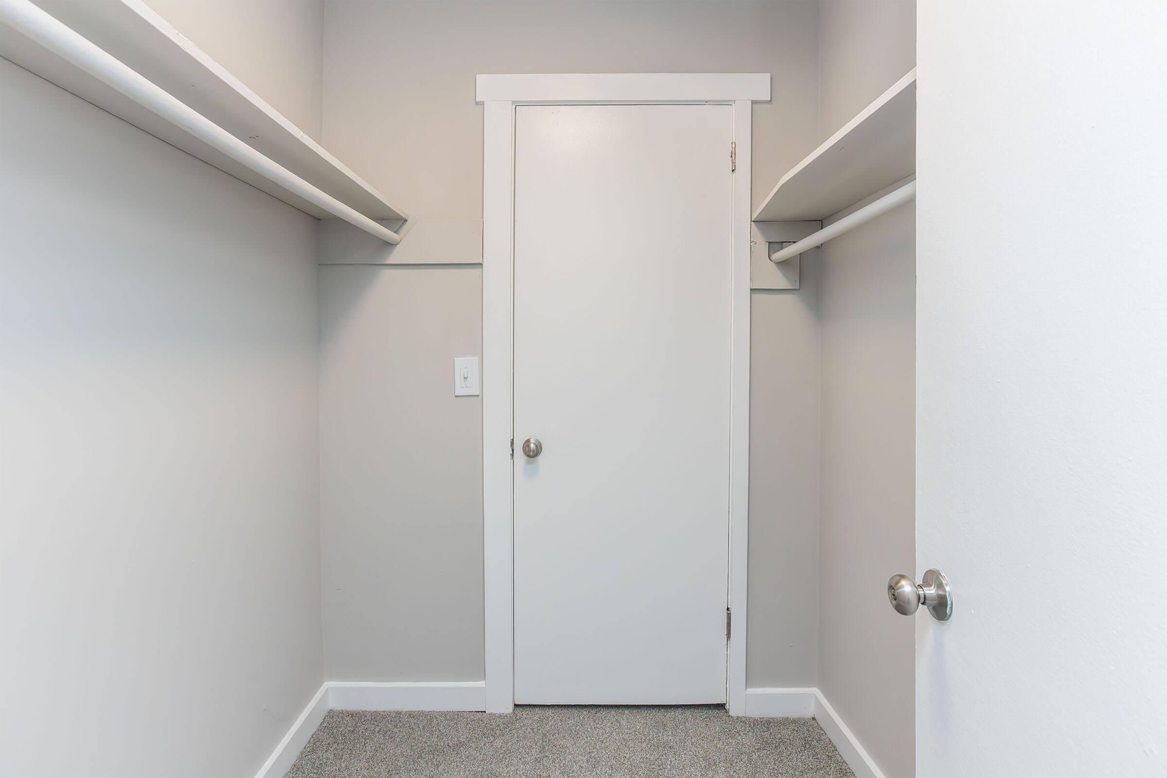 Empty closet with light gray walls, a single white door, and two shelves on either side. The floor is carpeted in a light color.