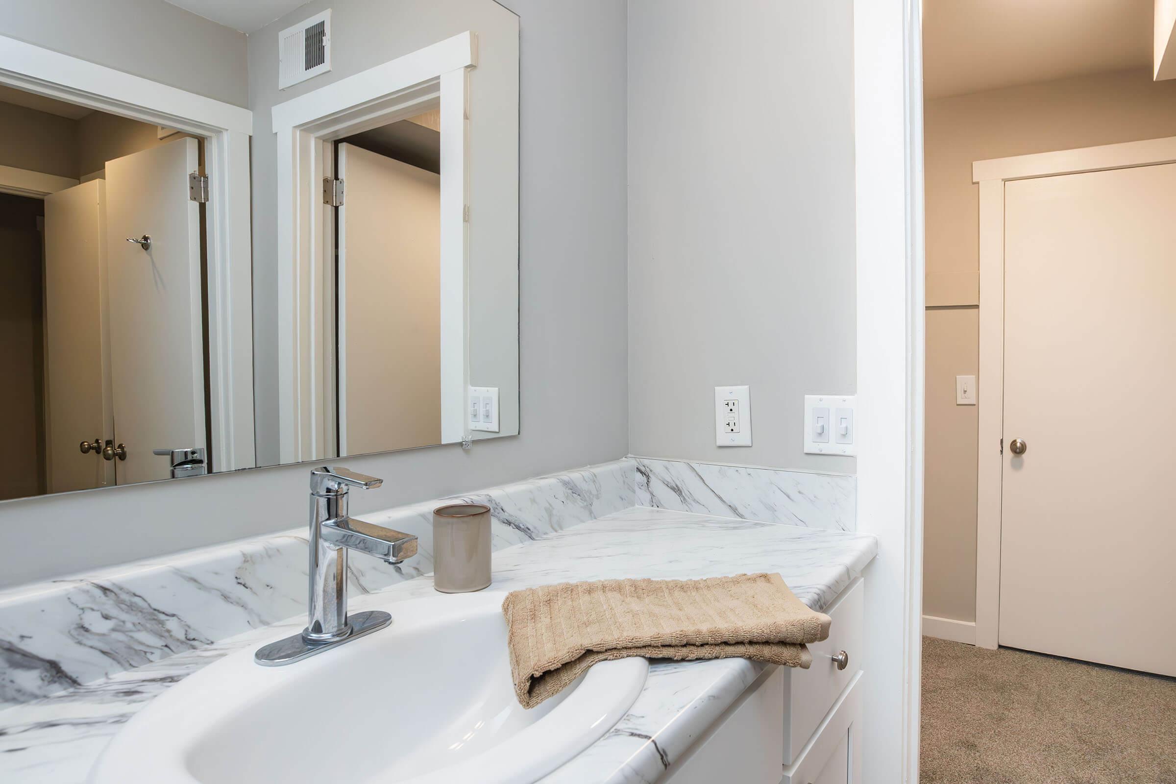A modern bathroom featuring a marble countertop with a white sink, a sleek faucet, a towel neatly placed beside it, and a lit candle. There are framed mirrors on the wall, light gray walls, and a door leading to another room in the background. The space is well-lit and stylish.