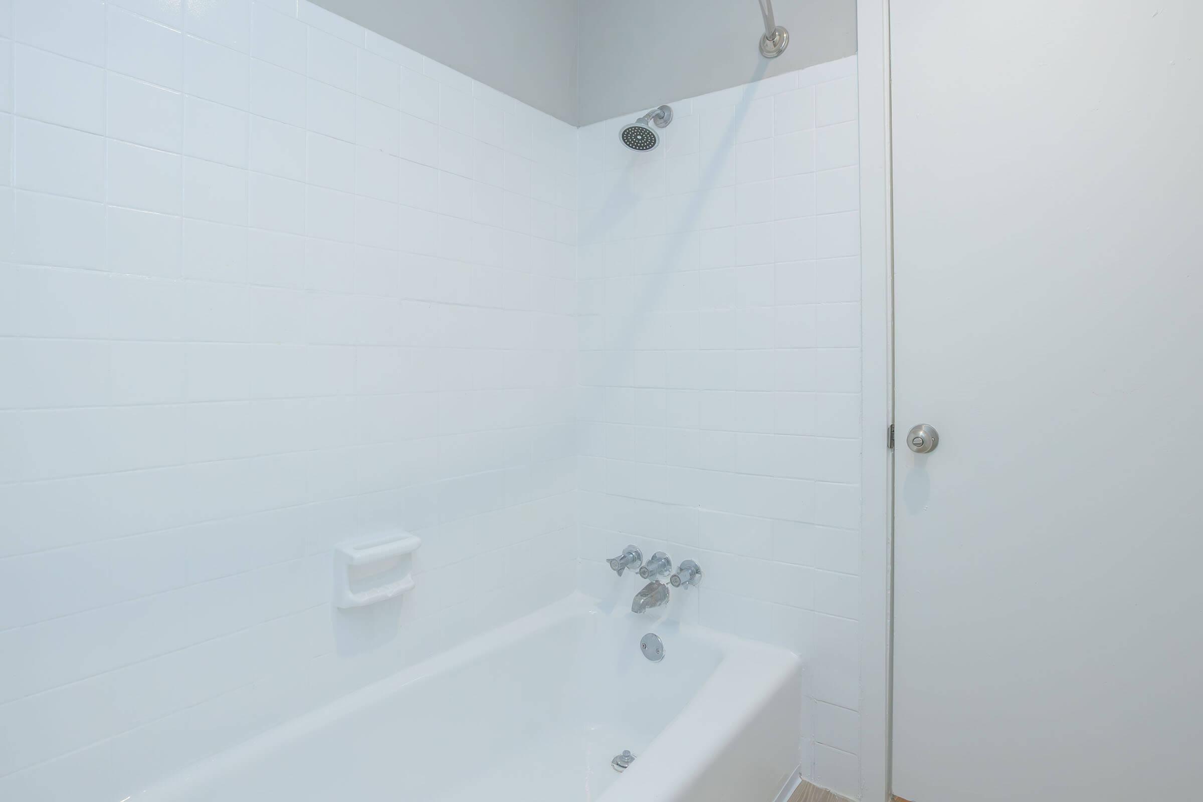 A clean, modern bathroom featuring a white tiled wall, a bathtub with silver fixtures, and a showerhead. A plain white door is partially visible on the right side. The overall aesthetic is simple and bright, emphasizing a minimalist design.