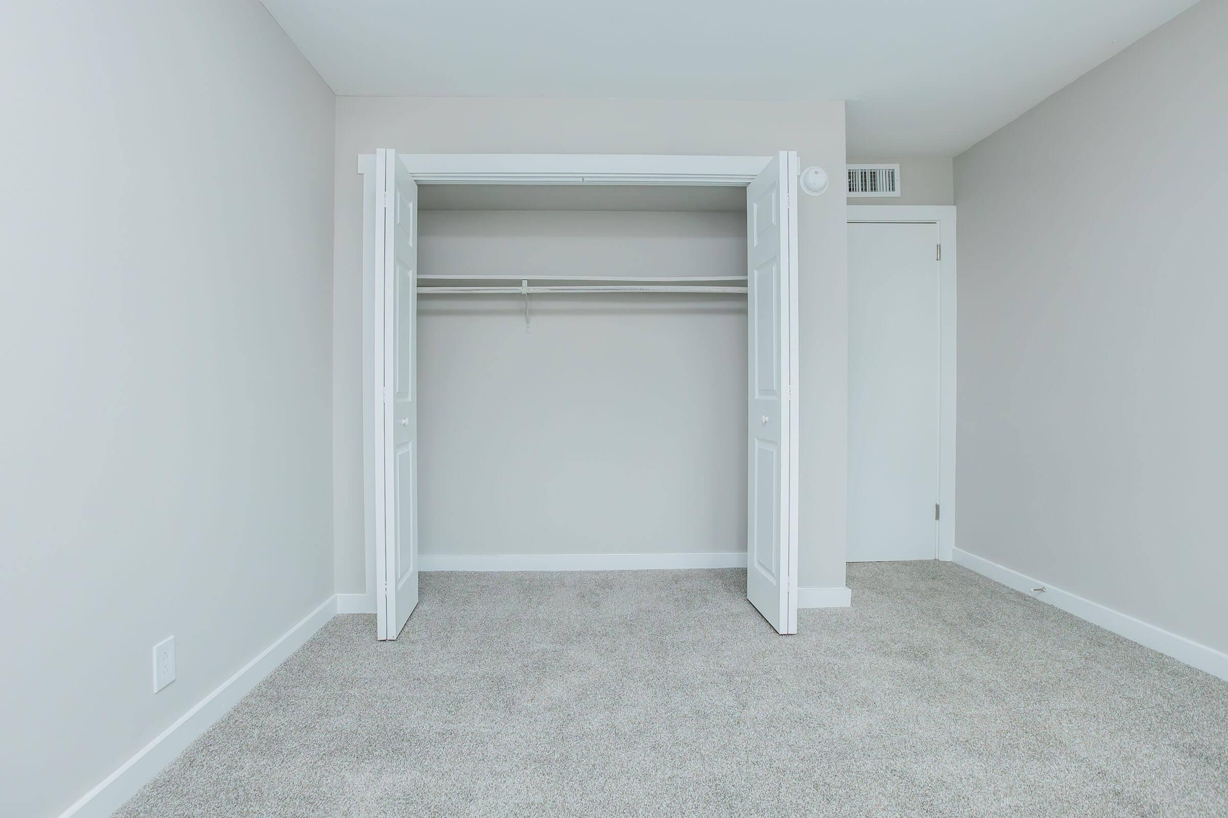 An empty closet with white double doors, partially open, revealing a single shelf inside. The walls are painted light gray, and the floor is covered with soft, light-colored carpet. A closed door is visible on the right side of the image. The overall impression is of a clean, minimalist space.