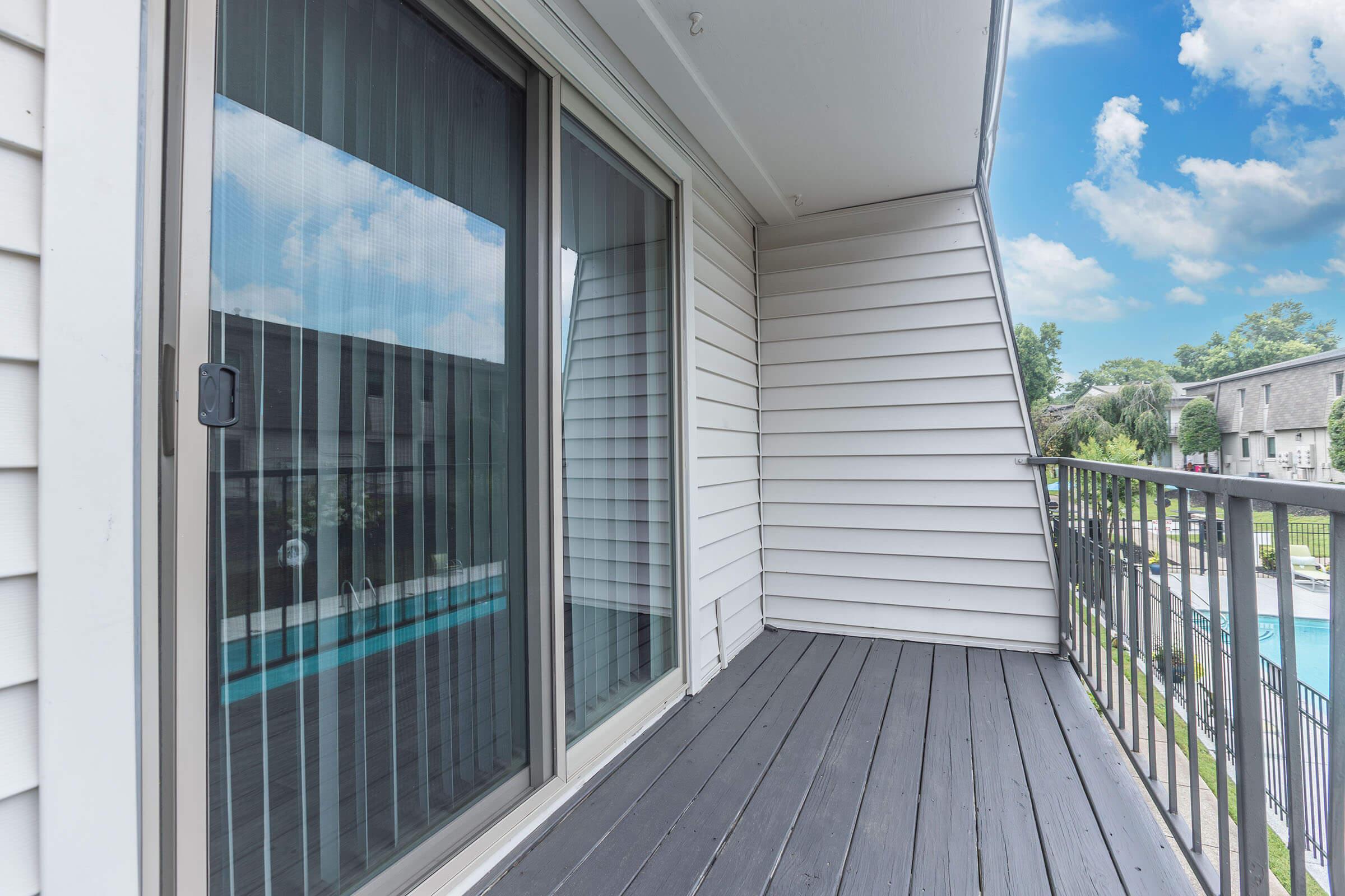 A balcony with a wooden floor and sliding glass doors reflecting the sky and surroundings. In the background, a glimpse of a pool and landscaped area can be seen through the glass. The walls are light-colored wood siding, and the atmosphere is bright and inviting.