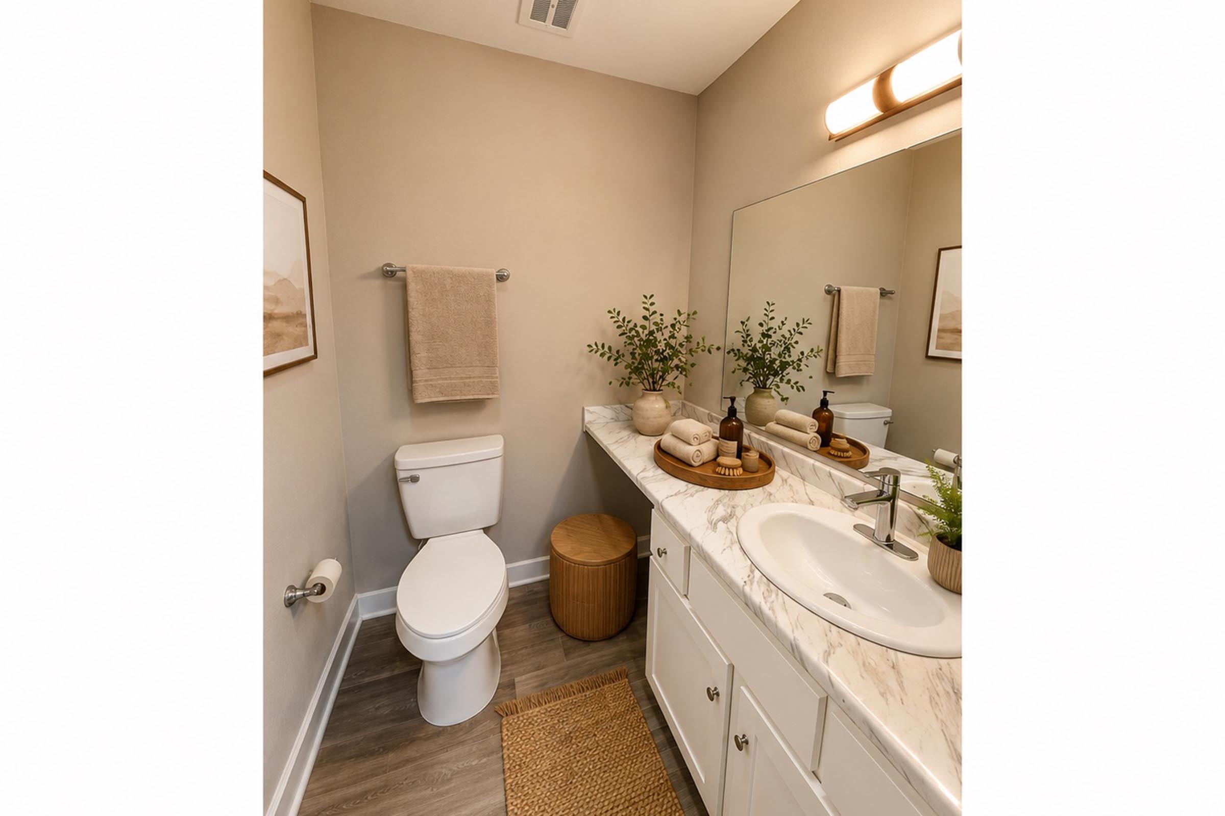 A modern bathroom featuring a white toilet, a marble countertop with a sink, and a large mirror above. The countertop is decorated with natural elements like a small plant and decorative items. Soft beige walls and a wooden basket add warmth, while a towel hangs on a rod and a rug lies on the floor.