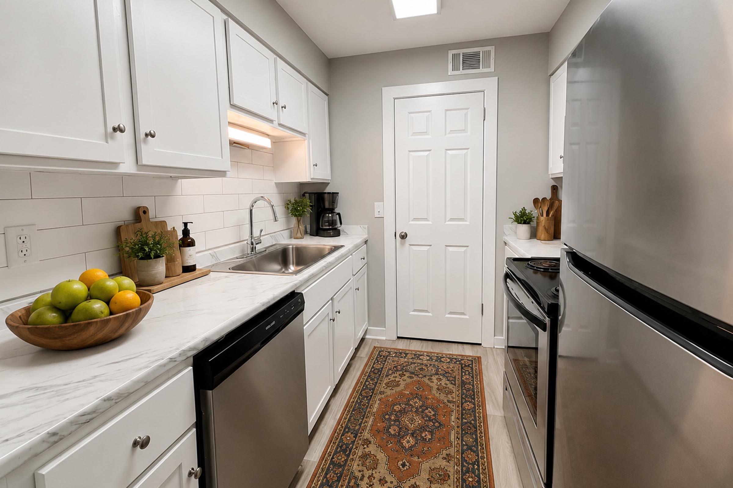 Bright and modern kitchen featuring white cabinets, a marble countertop, and stainless steel appliances. A bowl of green and yellow apples is on the counter next to a sink, with a coffee maker and potted plants nearby. The kitchen has a warm area rug, leading to a door with a white frame.