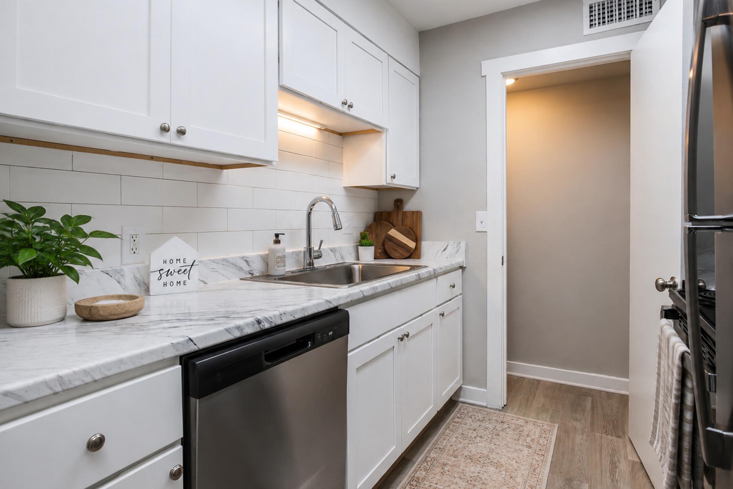 Modern kitchen featuring white cabinetry, a marble countertop, and stainless steel appliances. There is a small potted plant on the countertop, a wooden cutting board, and a decorative sign. The space is well-lit with a warm ambiance, and an open doorway leads to another room.