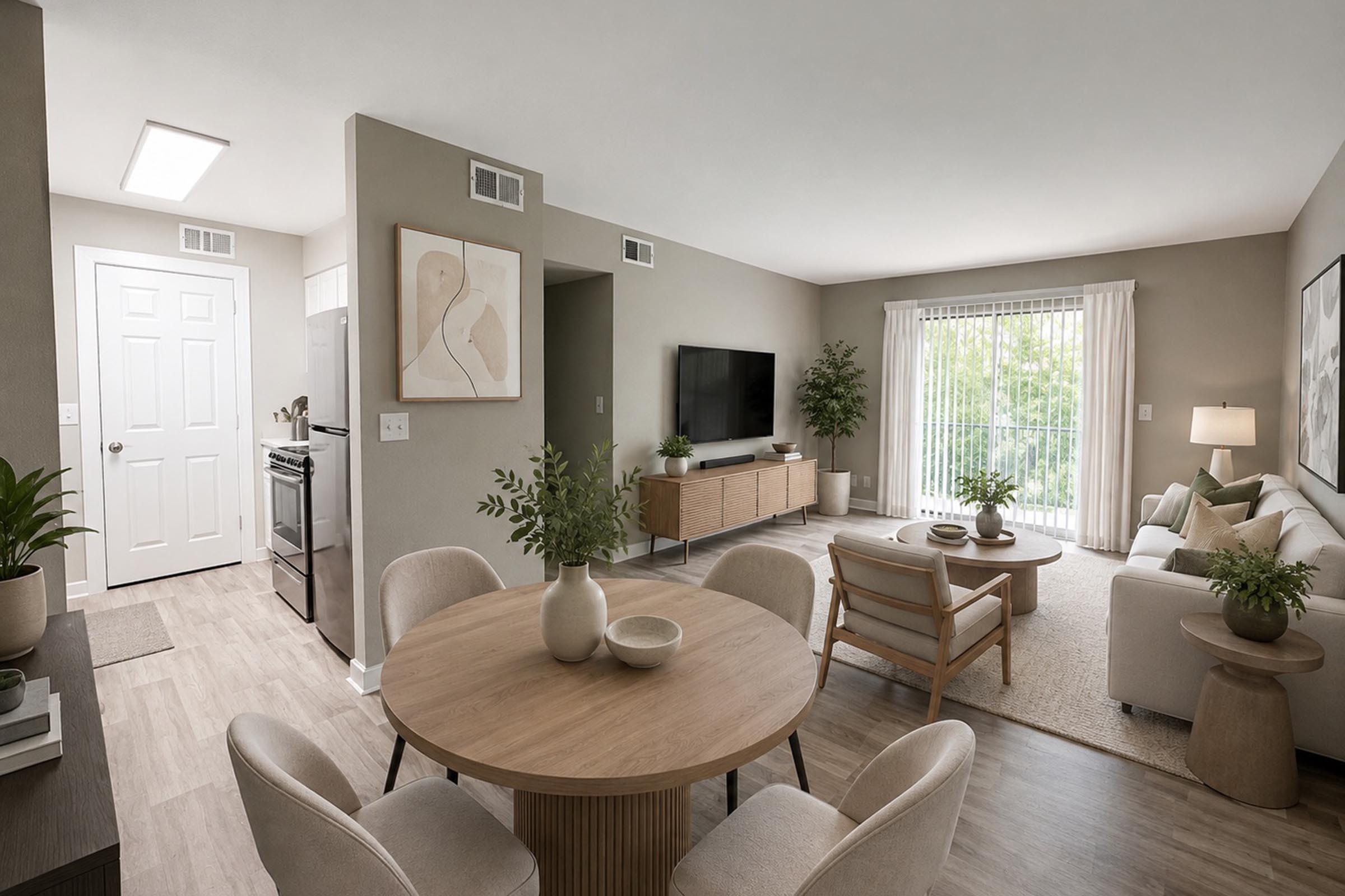 Bright and modern living area featuring a cozy sofa, a sleek TV unit, and a dining table with four chairs. The space has neutral walls and natural light flowing in through large windows, creating a welcoming atmosphere. Indoor plants add a touch of greenery to the decor.