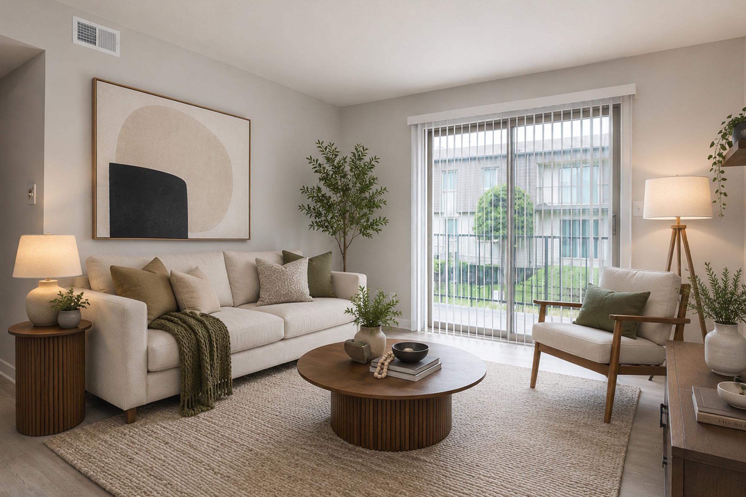 A cozy living room featuring a beige sofa adorned with green and tan pillows, a round wooden coffee table, and a light-colored area rug. To the right, there's a comfortable armchair and a floor lamp. Large windows with vertical blinds allow natural light to brighten the space, complemented by potted plants.