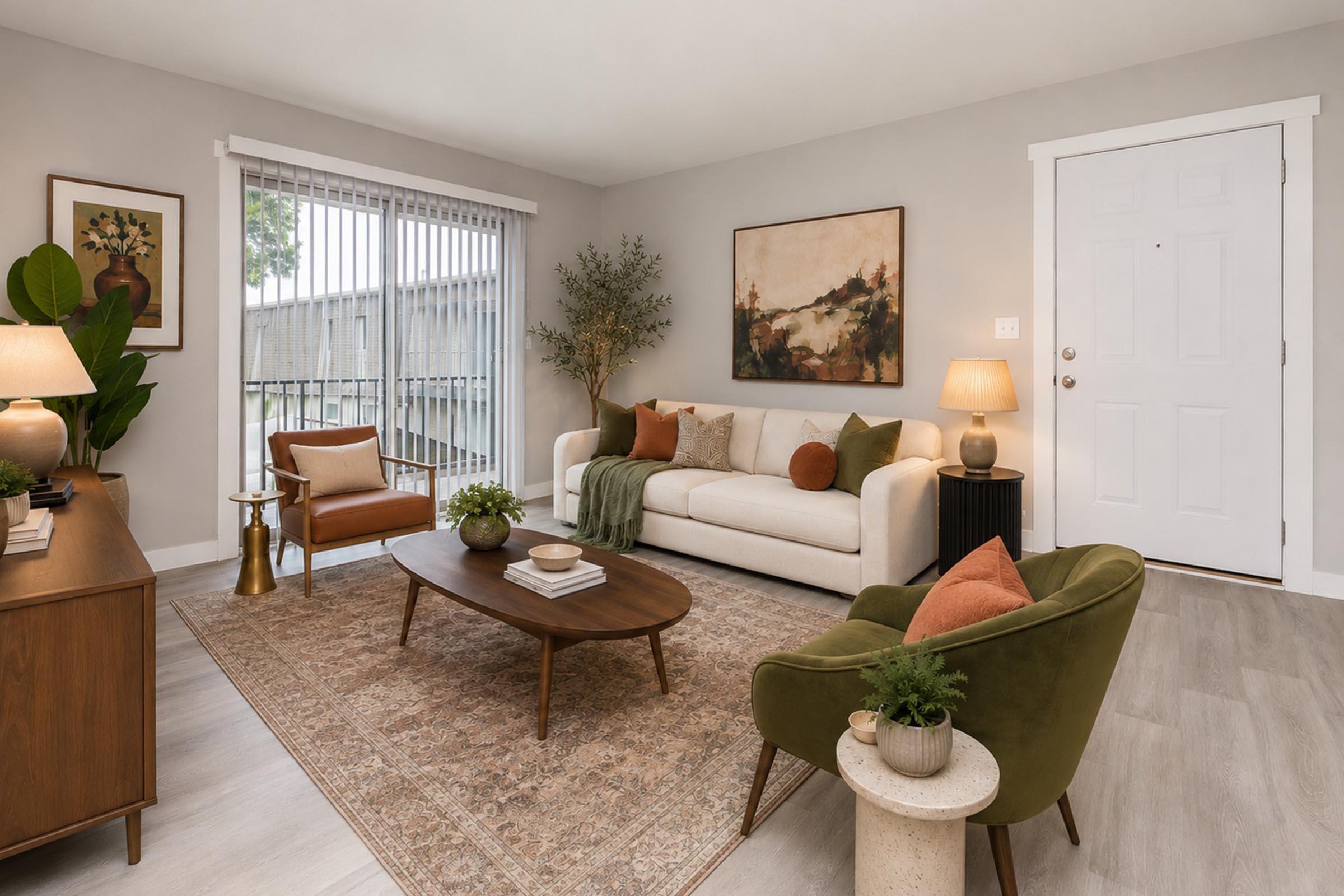 A cozy living room featuring a white sofa with green and orange accents, a round coffee table, and a stylish rug. There are two plants, a wooden sideboard, and warm lighting from table lamps. A large window with blinds lets in natural light, and a door leads outside. The walls are painted in a soft gray tone.