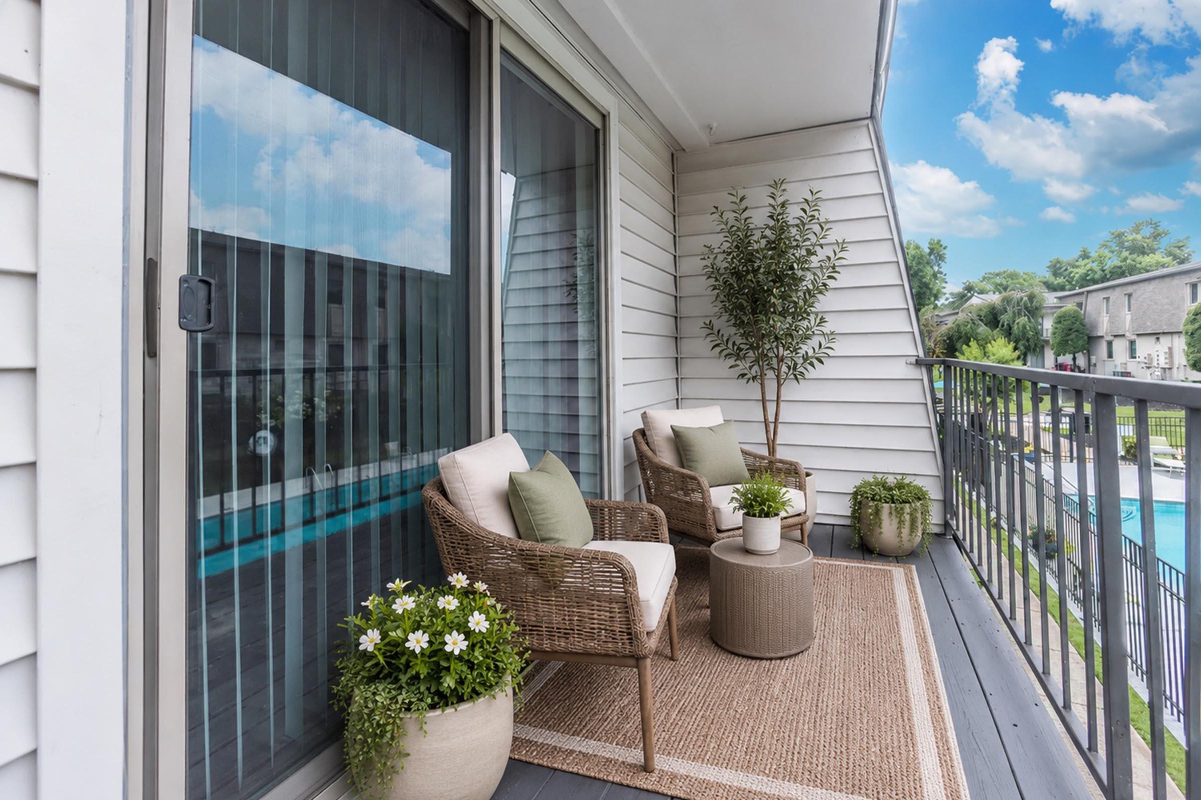 A cozy balcony with two wicker chairs and green cushions, adorned with potted plants and a rug. The backdrop features a view of a swimming pool and lush landscaping under a blue sky with clouds, creating a serene outdoor space for relaxation.