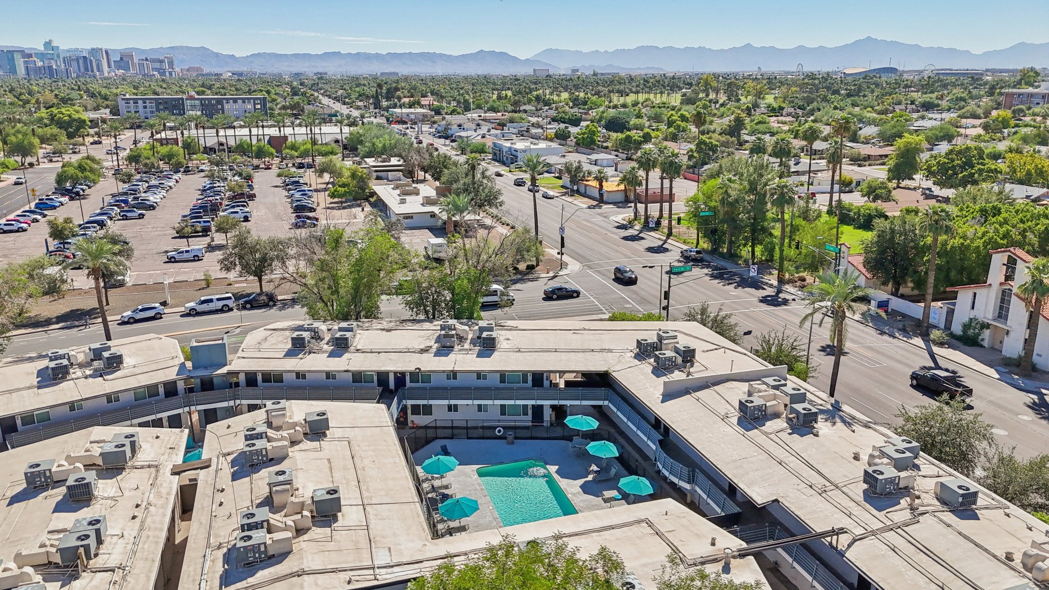 Aerial view of a neighborhood featuring a swimming pool surrounded by buildings. The image shows parked cars in a lot, lined streets, and mountains in the distance under clear skies. Lush greenery enhances the residential area, contributing to a vibrant atmosphere.