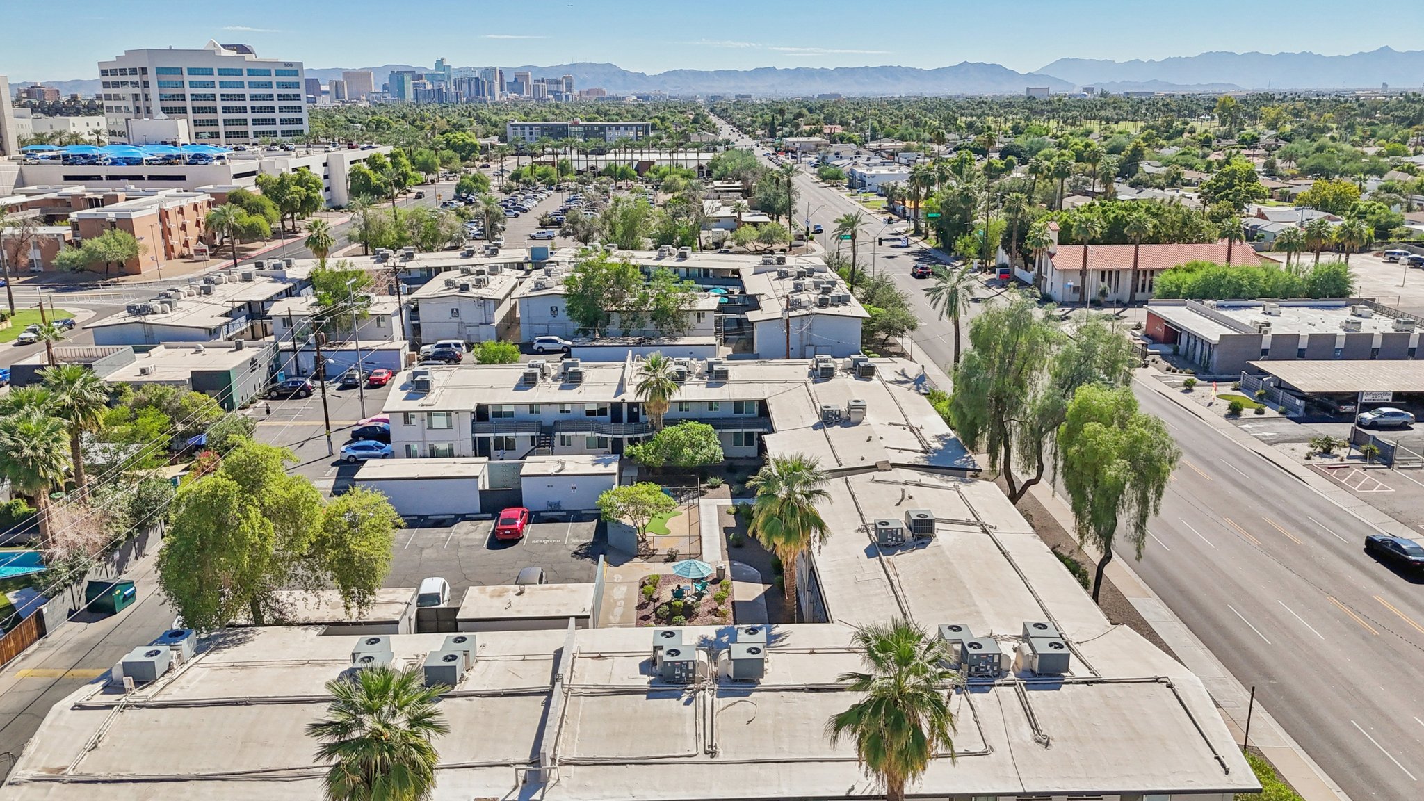 Aerial view of a suburban area featuring low-rise buildings with flat rooftops, palm trees, and parked cars. In the background, a skyline of a city is visible along with mountains in the distance. The scene captures a mix of residential and commercial structures, showcasing the greenery of the area.