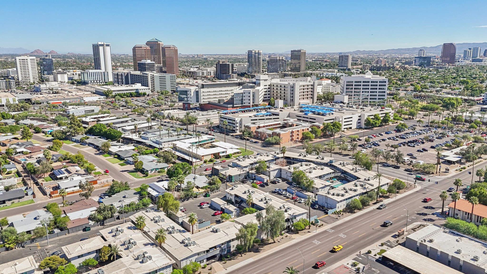 Aerial view of a cityscape featuring a mix of residential and commercial buildings. In the foreground, there are low-rise structures and a busy road with parked cars. In the background, taller skyscrapers rise against a clear blue sky, with mountains visible in the distance, showcasing a vibrant urban environment.