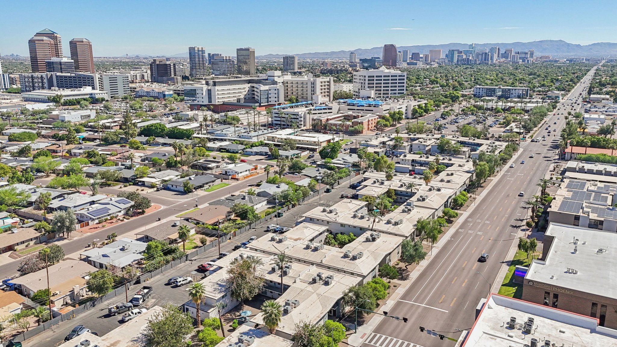 Aerial view of a sprawling urban landscape featuring a mix of residential and commercial buildings. Roads are lined with greenery, and taller buildings stand in the background against a clear blue sky. The city skyline and mountain range are visible in the distance. Traffic is present on the main road.