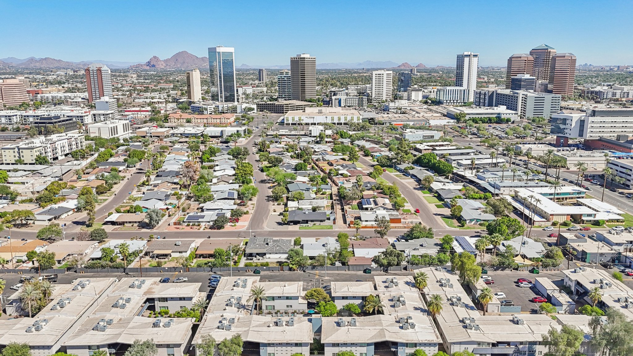 Aerial view of a cityscape featuring a mix of residential neighborhoods and high-rise buildings. In the foreground, there are low-rise housing developments, while the background showcases modern skyscrapers and mountainous terrain. Clear blue sky enhances the vibrant city setting.