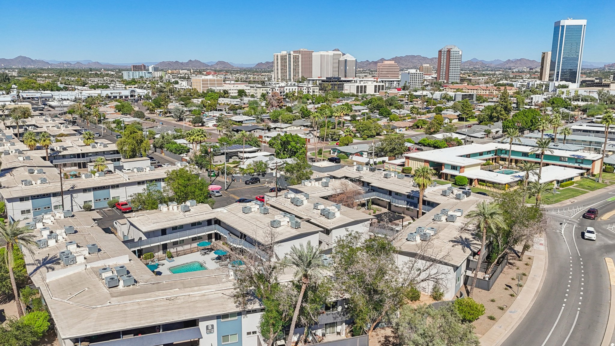 Aerial view of a city landscape featuring residential buildings and a few scattered palm trees. In the background, there are high-rise buildings and mountains. The scene showcases a clear blue sky with bright sunlight illuminating the area, highlighting the contrast between urban and natural elements.