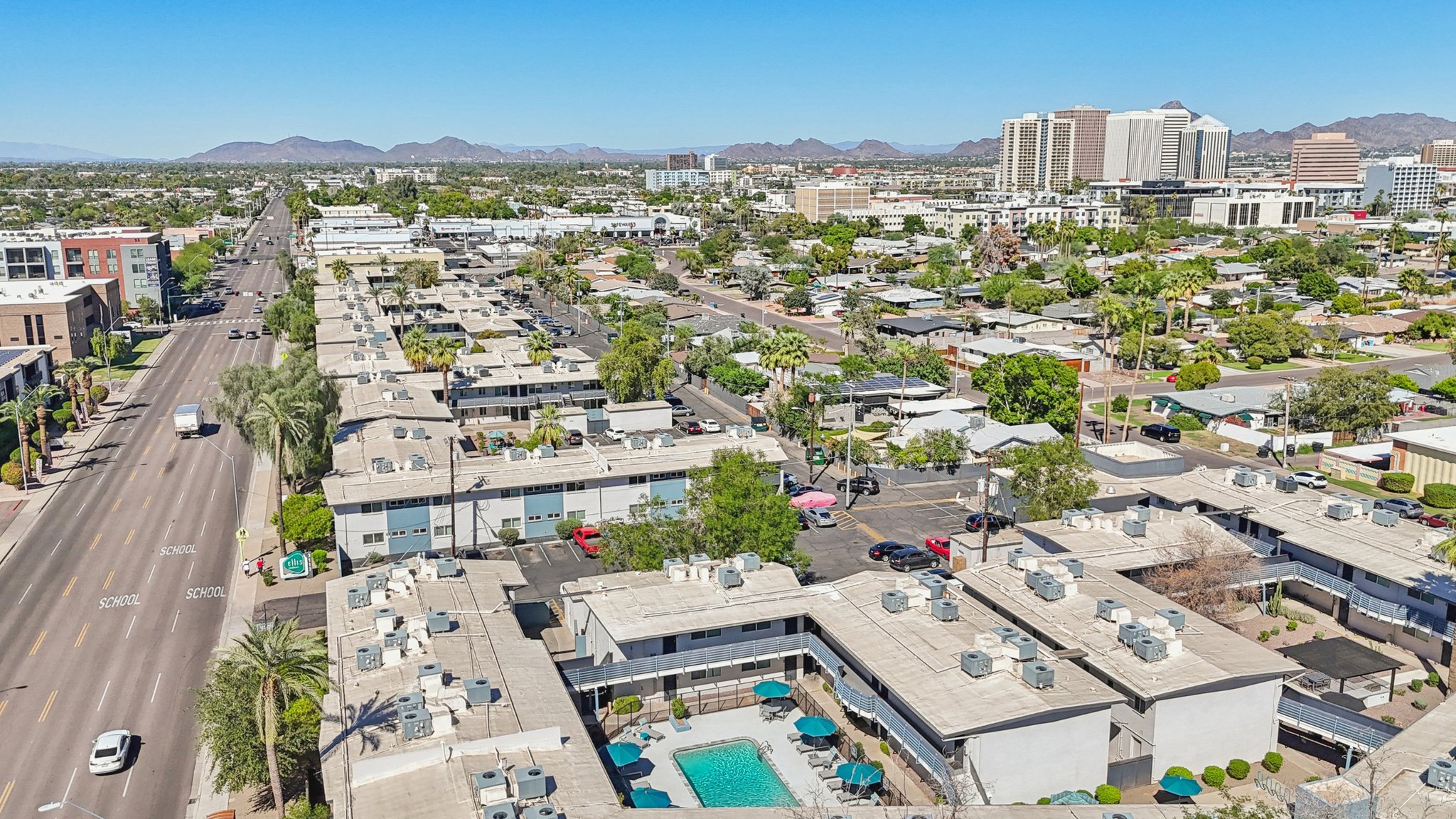Aerial view of a suburban area featuring low-rise buildings, a pool in the foreground, and a busy street lined with cars. In the distance, mountains are visible, along with taller buildings indicating an urban setting. Clear blue sky enhances the overall bright atmosphere of the scene.