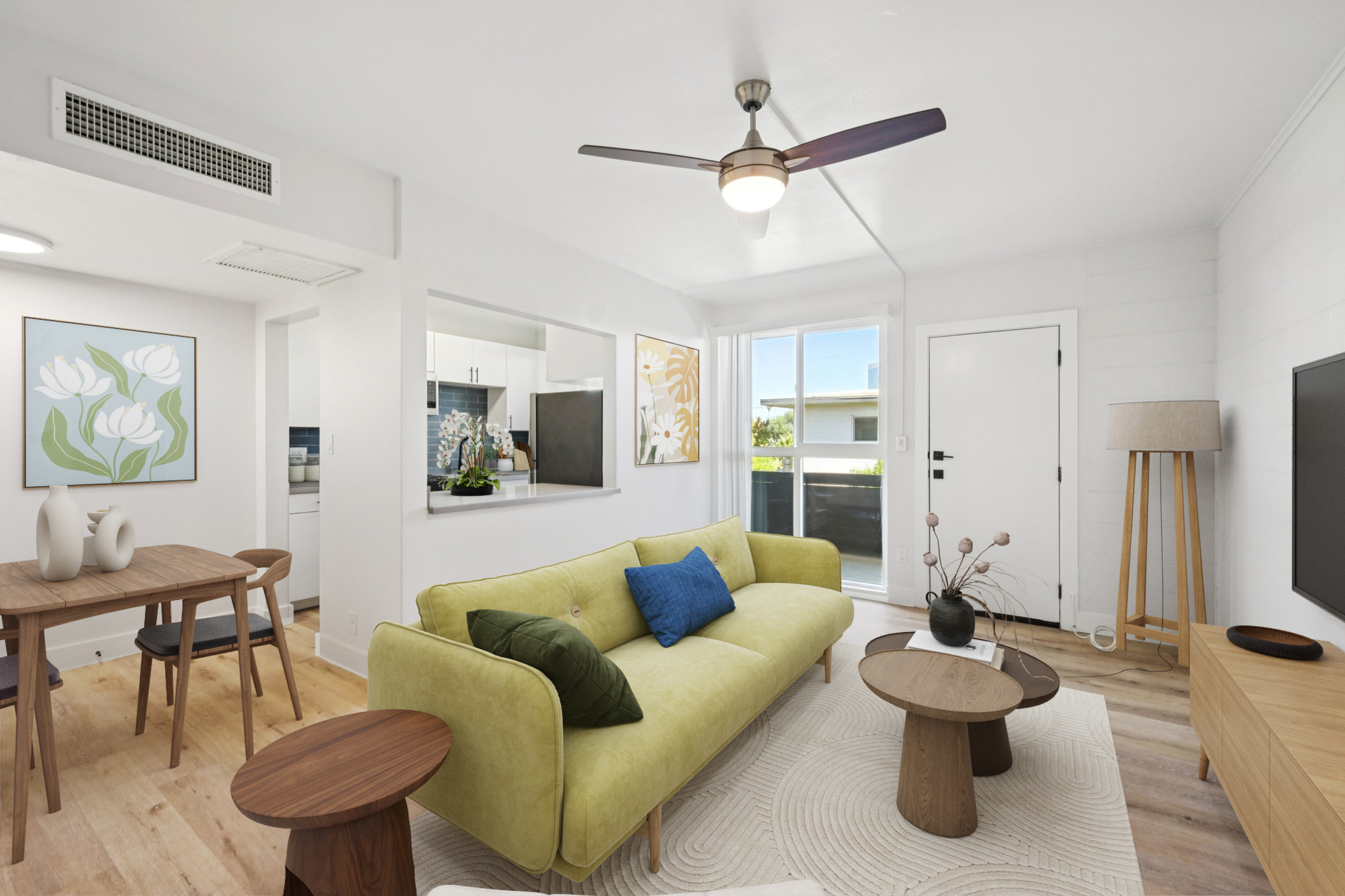 Bright, modern living room featuring a light green sofa with a blue cushion, a round wooden coffee table, and a dining area with a wooden table and chairs. Wall art and a light wood floor enhance the space, which has large windows and a fan overhead for a fresh, airy atmosphere.