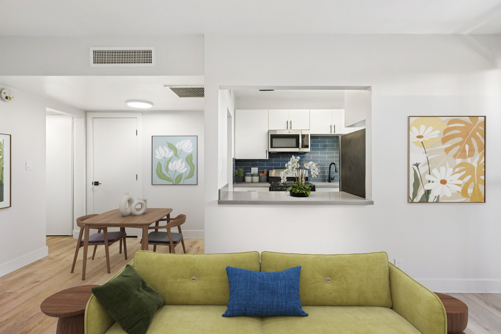 A modern living area featuring a bright green sofa with a blue cushion, adjacent to a small dining table. The kitchen is visible in the background, showcasing white cabinetry and blue tile. Two floral artworks decorate the walls, adding a touch of color to the space, which is well-lit and has wooden flooring.