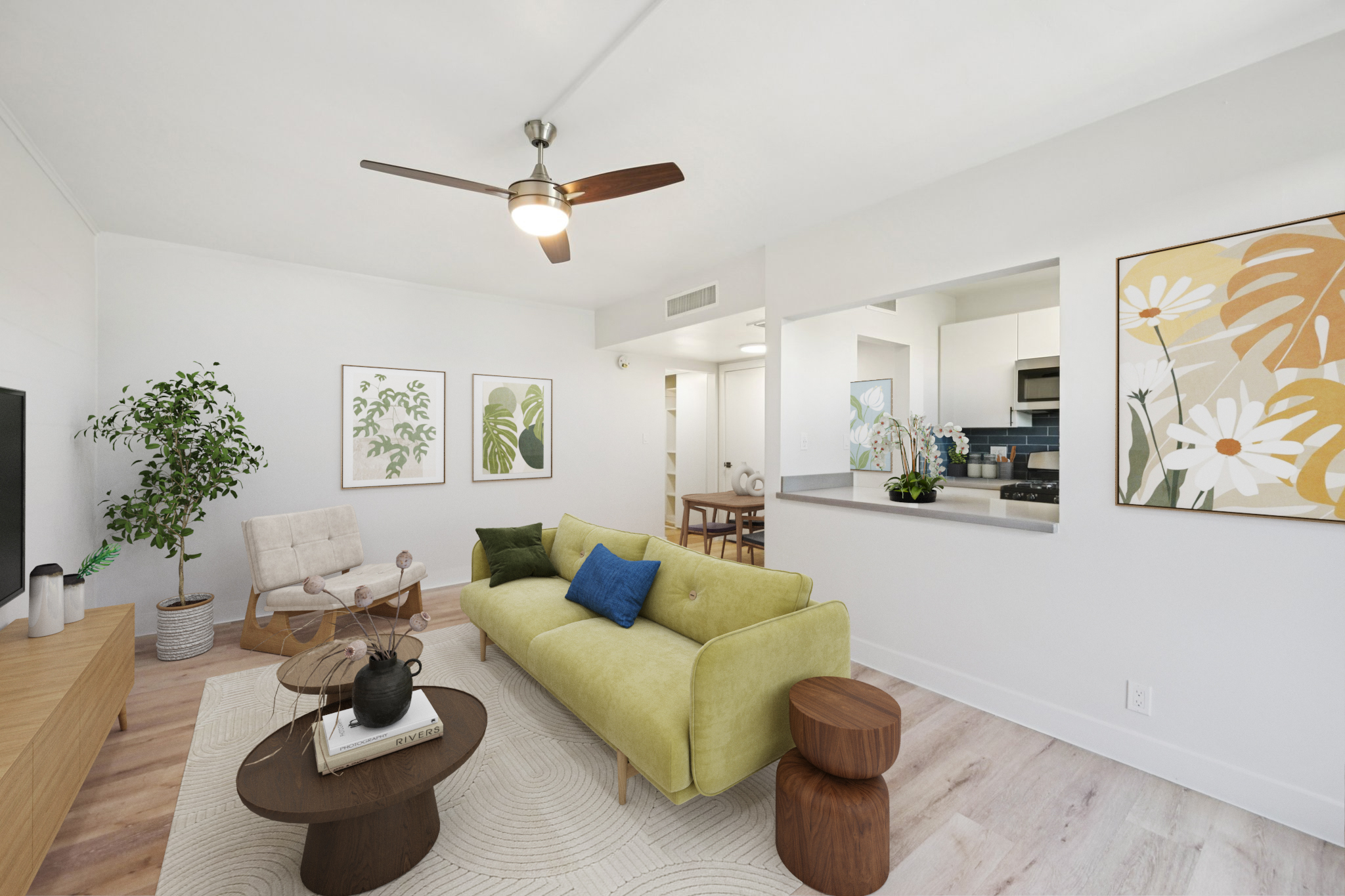 A modern living room featuring a green sofa with blue pillows, a wooden side table, and a decorative plant. A lighted ceiling fan hangs above, while a large TV is mounted on the wall. The room has artwork depicting floral designs and an open kitchen area in the background, creating a bright and inviting atmosphere.