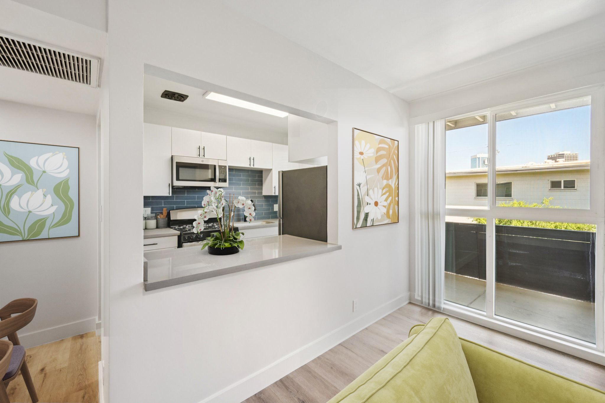 Bright and modern apartment interior featuring an open kitchen with white cabinetry, stainless steel appliances, and a blue tile backsplash. A cozy living area with a green sofa, decorative wall art of flowers, and large windows allowing natural light, creating a welcoming atmosphere.