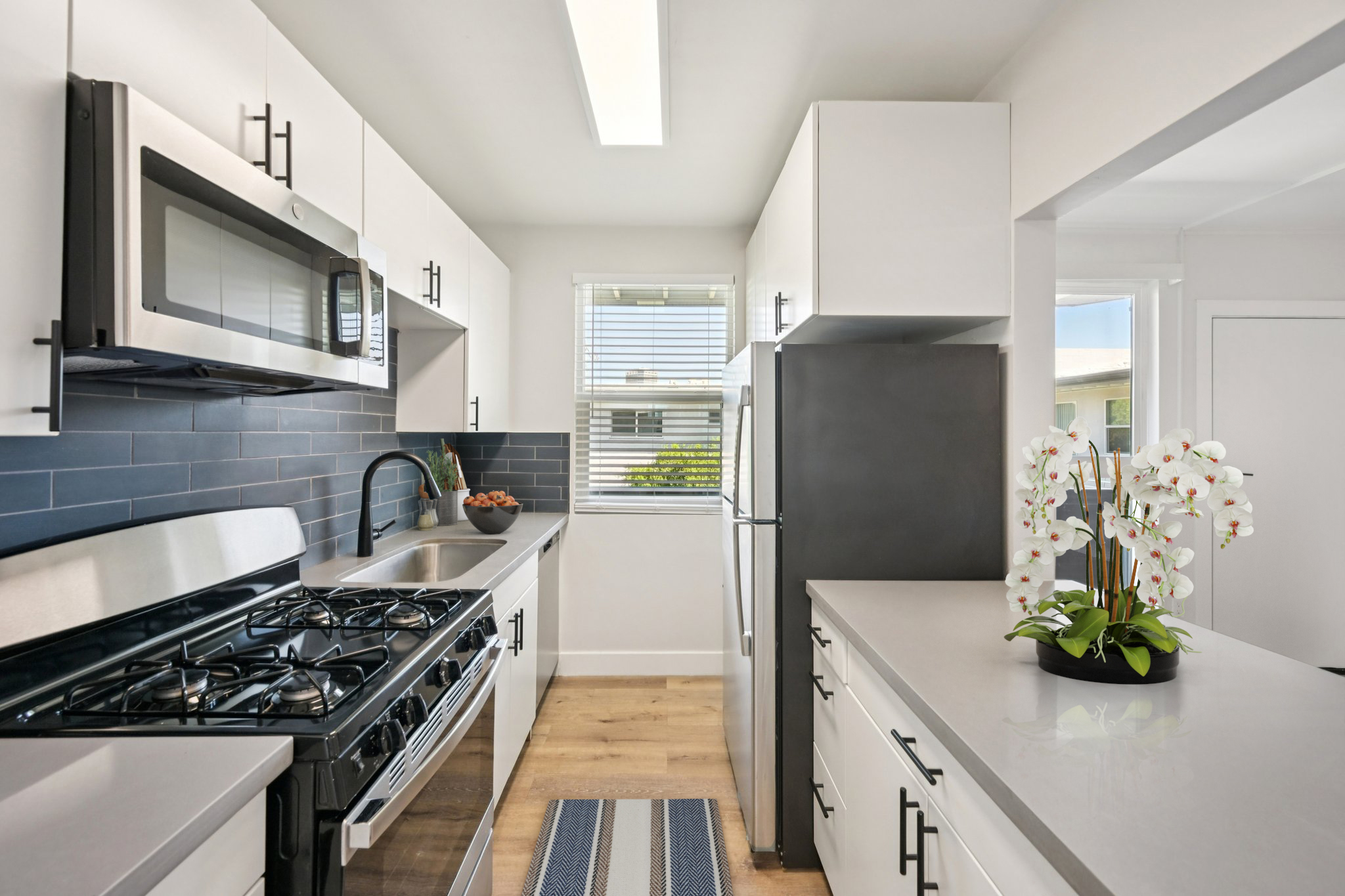 Modern kitchen featuring stainless steel appliances, including a gas stove and microwave. The cabinetry is white with sleek black hardware, and a built-in sink overlooks a window with natural light. A black refrigerator stands opposite, and a small island with a planter of white orchids adds a touch of elegance.