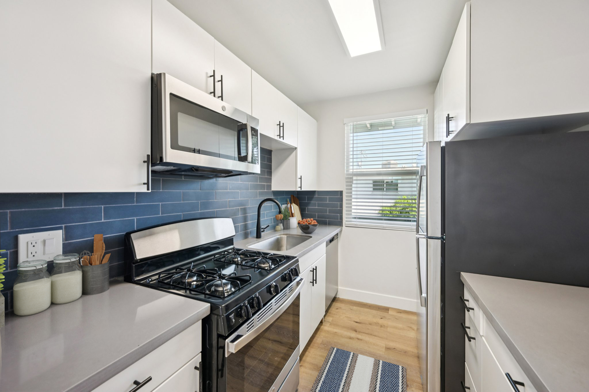 Modern kitchen featuring white cabinetry, stainless steel appliances, and a gray countertop. It includes a gas stove, microwave, and a black refrigerator, with a dark blue tile backsplash and a window providing natural light. A striped rug adds a touch of style to the wooden floor.