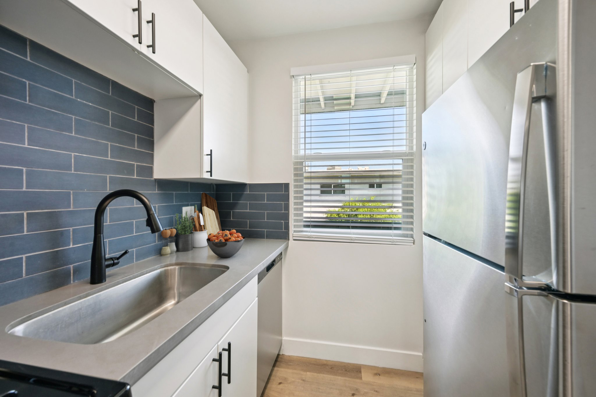 Modern kitchen with sleek white cabinetry, blue tiled backsplash, and stainless steel appliances. A window allows natural light to illuminate the space. A bowl of fruit sits on the counter, adding a pop of color to the minimalist design. The kitchen features a contemporary faucet and a clean, organized layout.