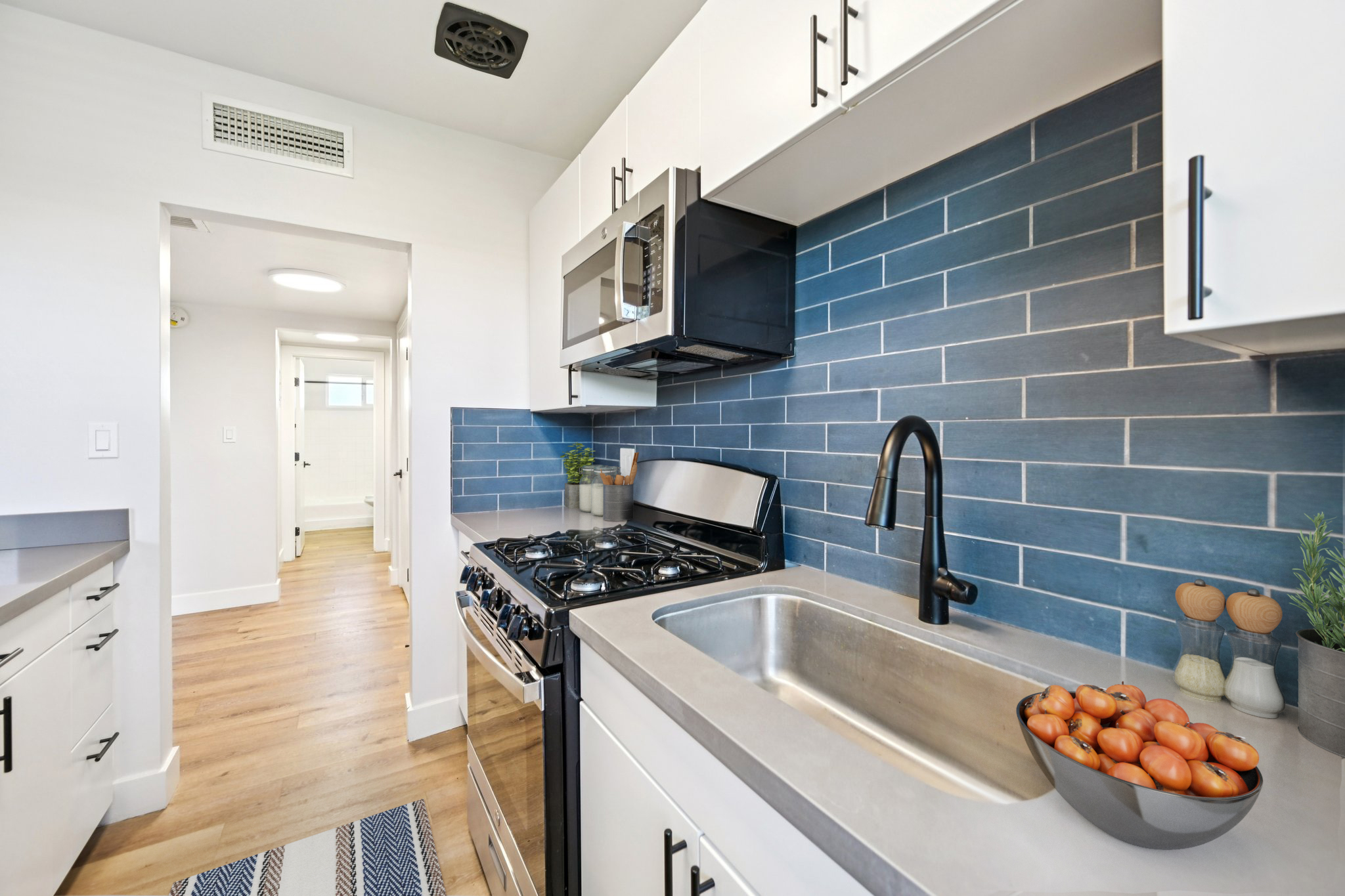 A modern kitchen featuring white cabinets, a stainless steel oven and microwave, a black faucet, and a gray countertop. The backsplash is composed of blue tiles, and a bowl of tomatoes sits on the counter. A corridor with a door can be seen in the background, enhancing the open feel of the space.