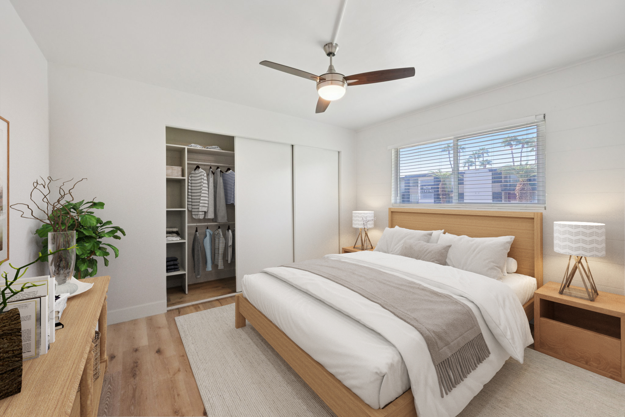 A modern bedroom featuring a queen-size bed with light bedding and a textured throw. Two bedside tables with lamps are on either side. A ceiling fan hangs above, and a spacious closet with sliding doors is visible in the background, showcasing organized clothing. Natural light streams in through a window.