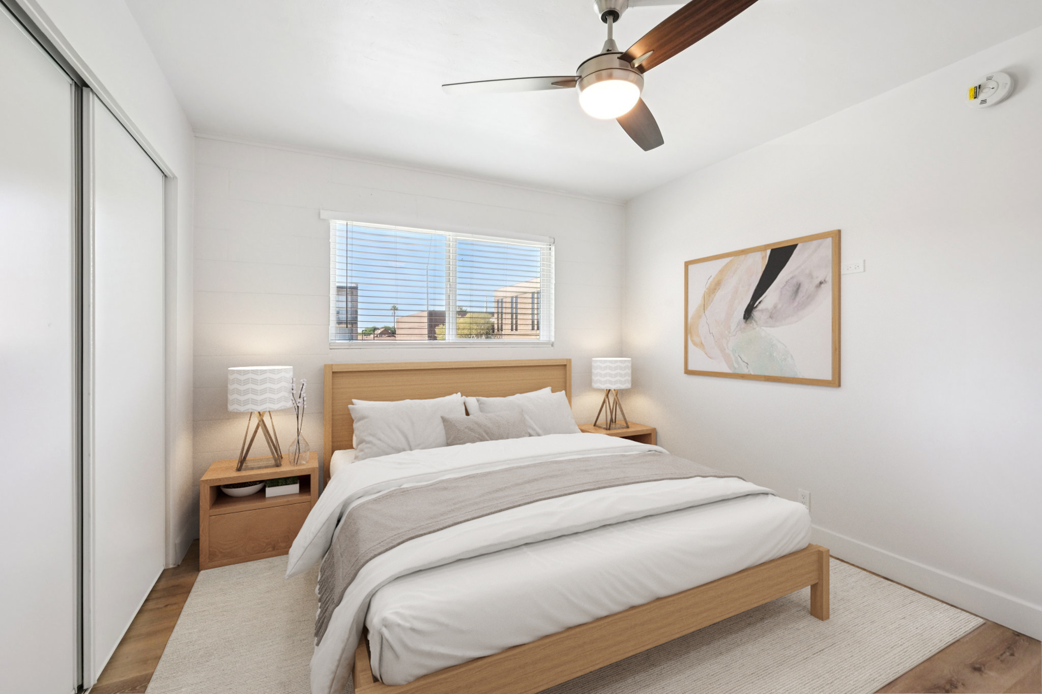 A cozy, modern bedroom featuring a wooden bed frame with white bedding, two bedside lamps on matching wooden nightstands, and a large window allowing natural light. The room has light-colored walls, a simple area rug, and a piece of abstract artwork on the wall.