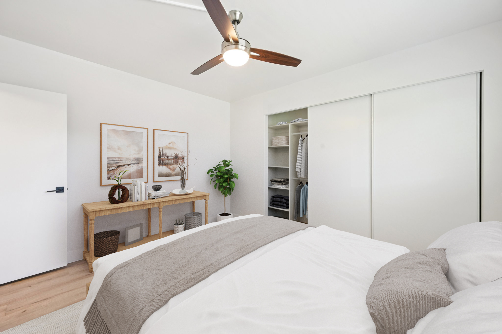 A modern bedroom featuring a light-colored wall, a cozy bed with gray and white bedding, and a wooden table adorned with decorative items. A closet with sliding doors is visible, along with a potted plant in the corner. Two framed art pieces hang above the table, adding a touch of style to the space.