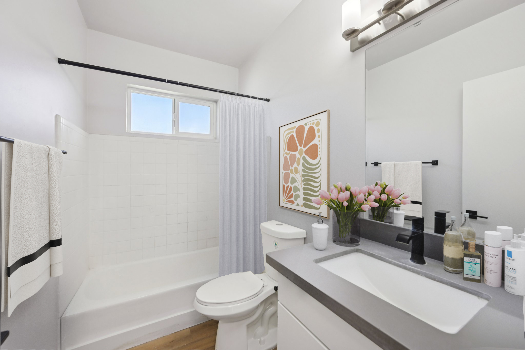 Bright and modern bathroom featuring a white bathtub with a shower curtain, a sleek vanity with a grey countertop, and various toiletries. Natural light enters through a window, complemented by a framed artwork and a vase of pink flowers on the counter. Clean, minimalist design with soft color palette.