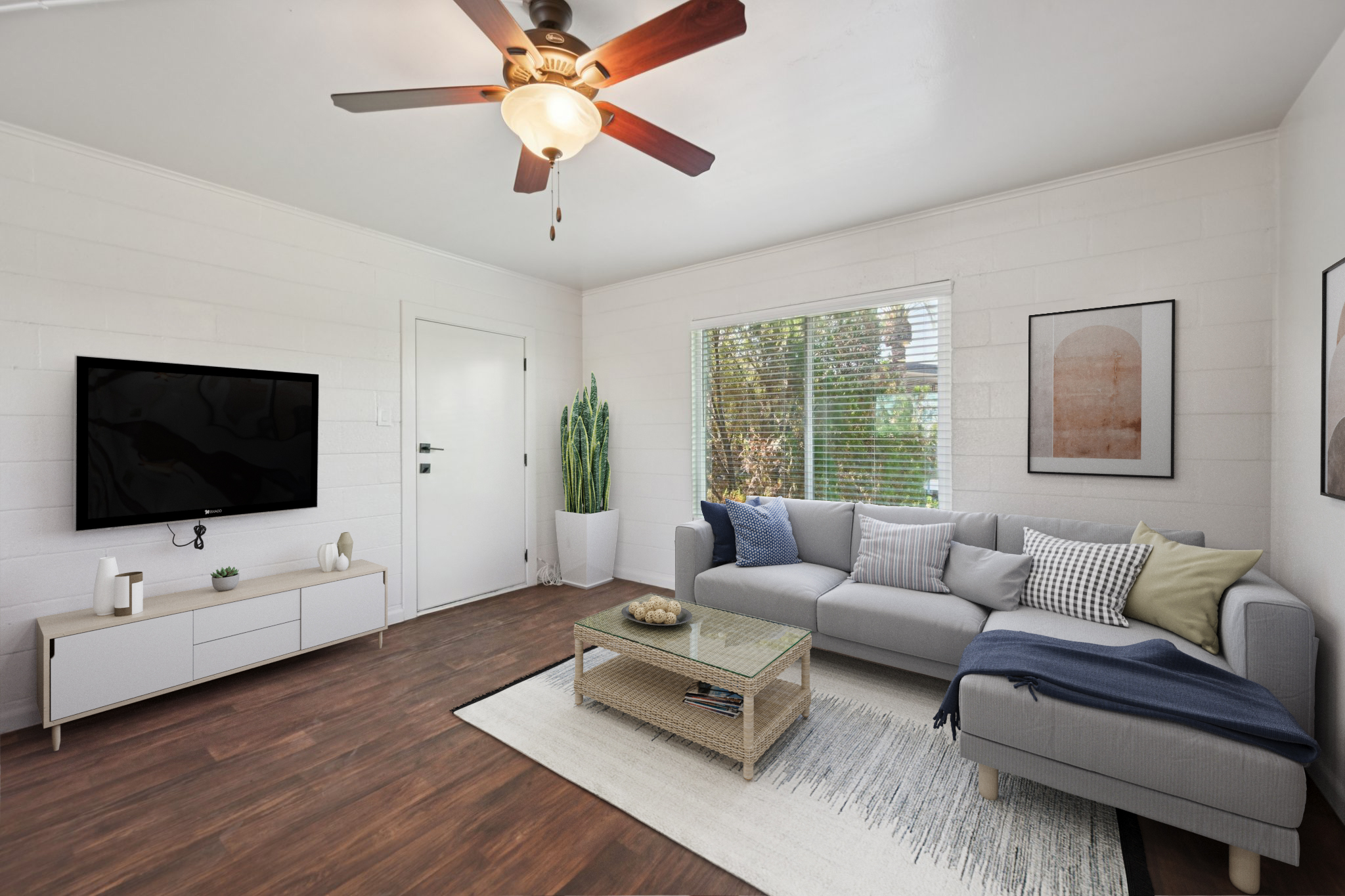 A modern living room featuring a light gray sectional sofa with colorful throw pillows, a glass coffee table on a patterned rug, and a flat-screen TV mounted on a white wall. Natural light streams through a large window, illuminating a tall indoor plant in the corner. A simple decorative vase and art pieces complete the cozy atmosphere.