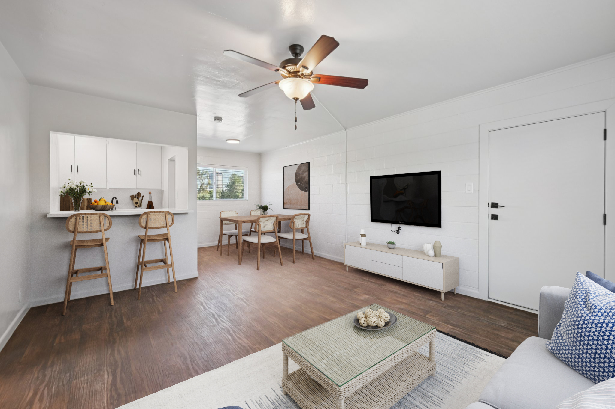 A modern living room with wooden flooring, featuring a white sofa with blue cushions, a coffee table with decorative items, and a wall-mounted television. There is a dining area with a light wood table and four chairs. The kitchen area is open with white cabinets and bar stools at the counter. Natural light enters through a window.