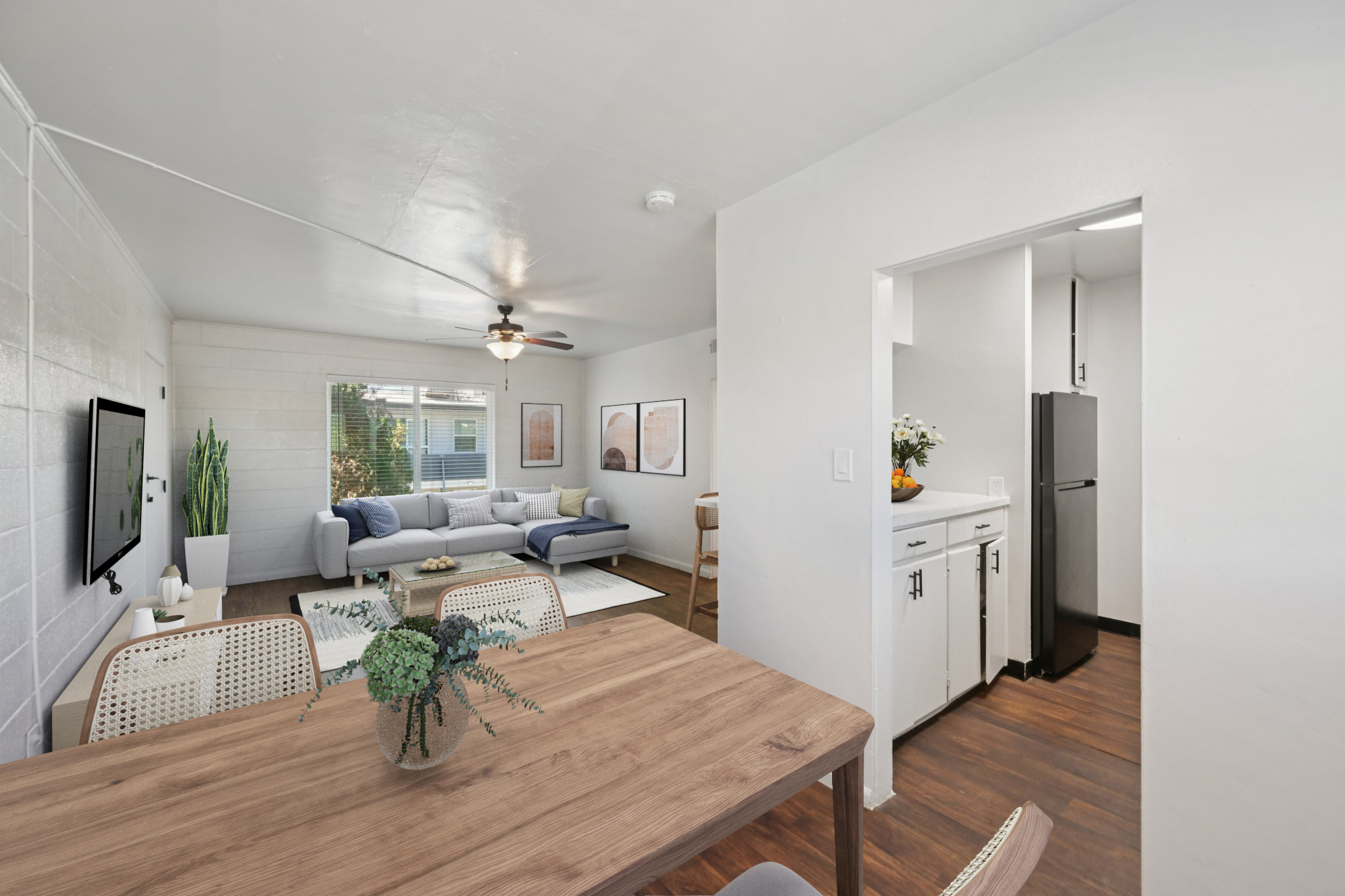 A bright and airy living space featuring a cozy gray sofa with decorative pillows, a wooden dining table with a centerpiece, and a modern kitchen visible in the background. Large windows allow natural light to flood the area, enhancing the welcoming atmosphere. Neutral colors and minimalist decor create a calm environment.