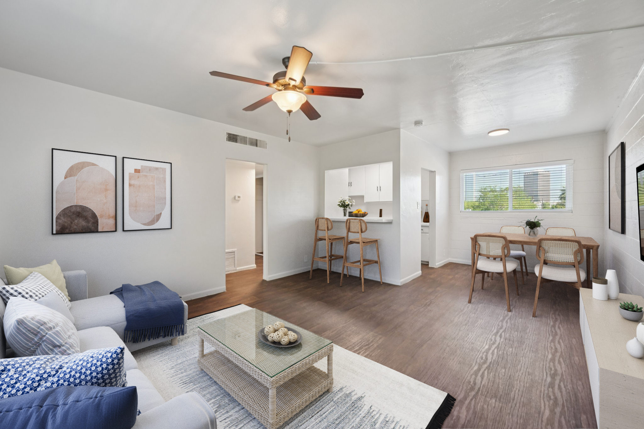 A bright and modern living room featuring a white sofa with decorative pillows, a coffee table with a decorative bowl, and two framed geometric art pieces on the wall. To the back, there is a dining area with a wooden table and chairs, and a small kitchen bar. Natural light streams in through the window.