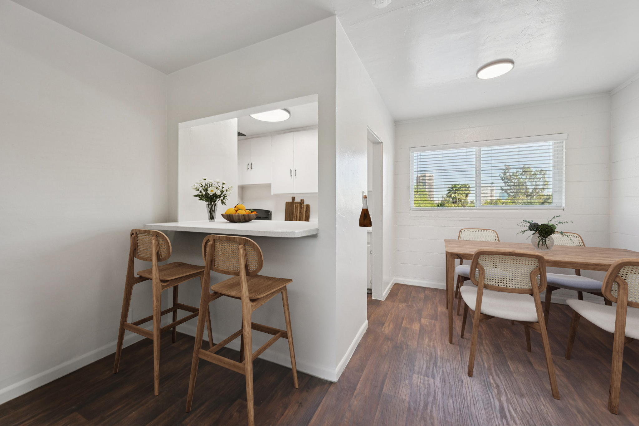 A modern kitchen and dining area with a white color scheme. There are two wooden bar stools at a small counter, and a simple dining table with chairs. Fresh flowers and fruit are on the counter, and a window lets in natural light, revealing greenery outside. The floor has a warm wood finish.