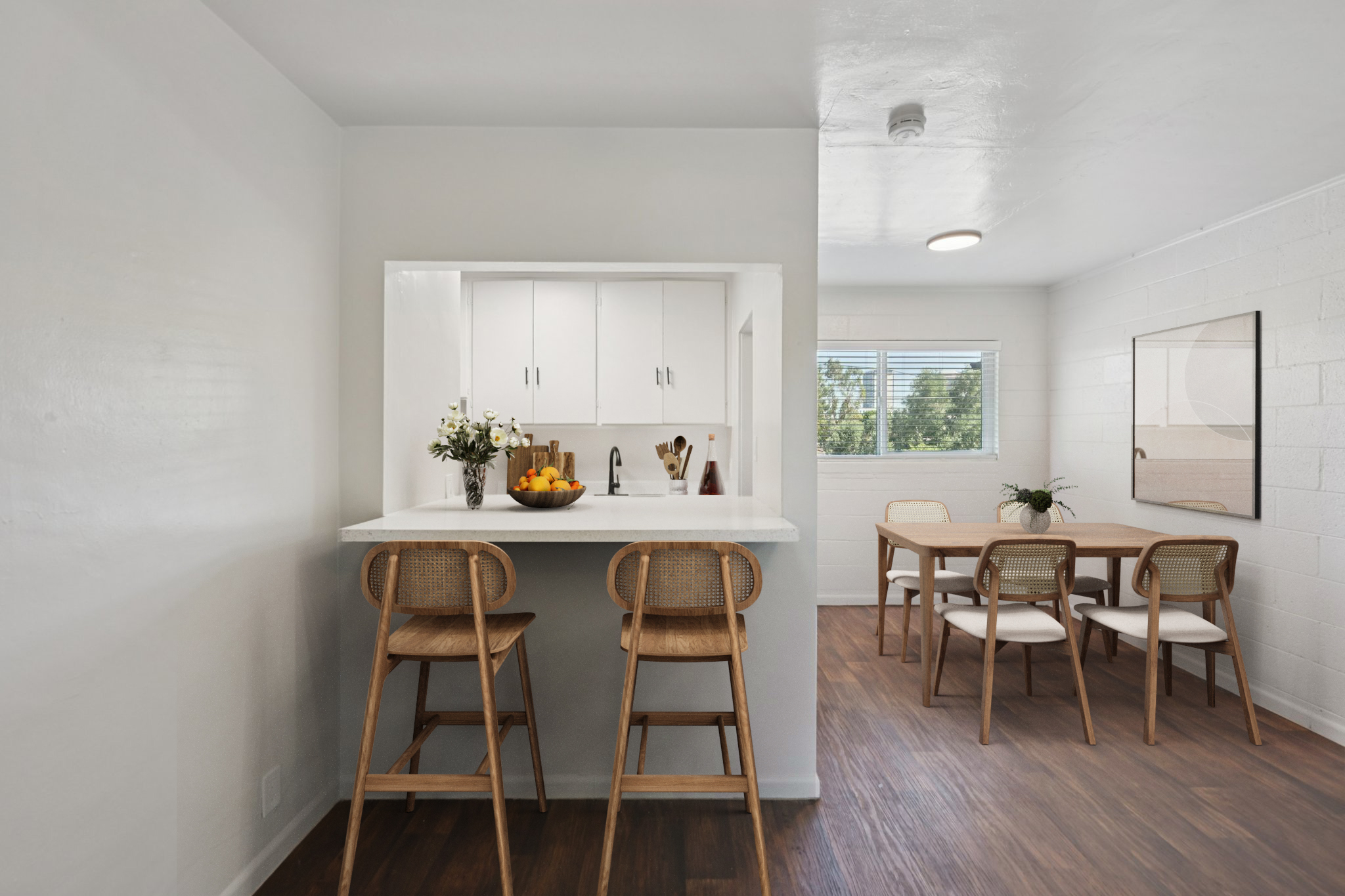 A modern kitchen area featuring a bar with three wooden stools, a bowl of fruit, and a small flower arrangement. Adjacent is a dining space with a wooden table and four chairs. The interior has a bright, minimalist design with white walls and wooden flooring, illuminated by natural light from windows.