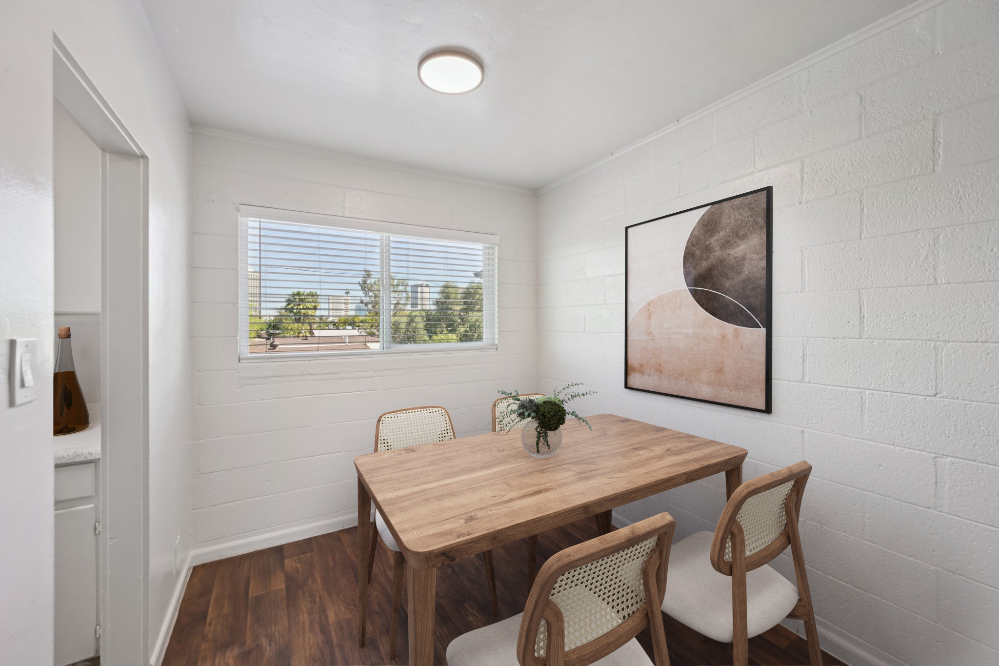 A bright, minimalistic dining area featuring a wooden table with three light-colored chairs. A potted plant sits on the table. The walls are painted white with a large, modern abstract art piece framed on one side. Natural light flows through a window, illuminating the space.