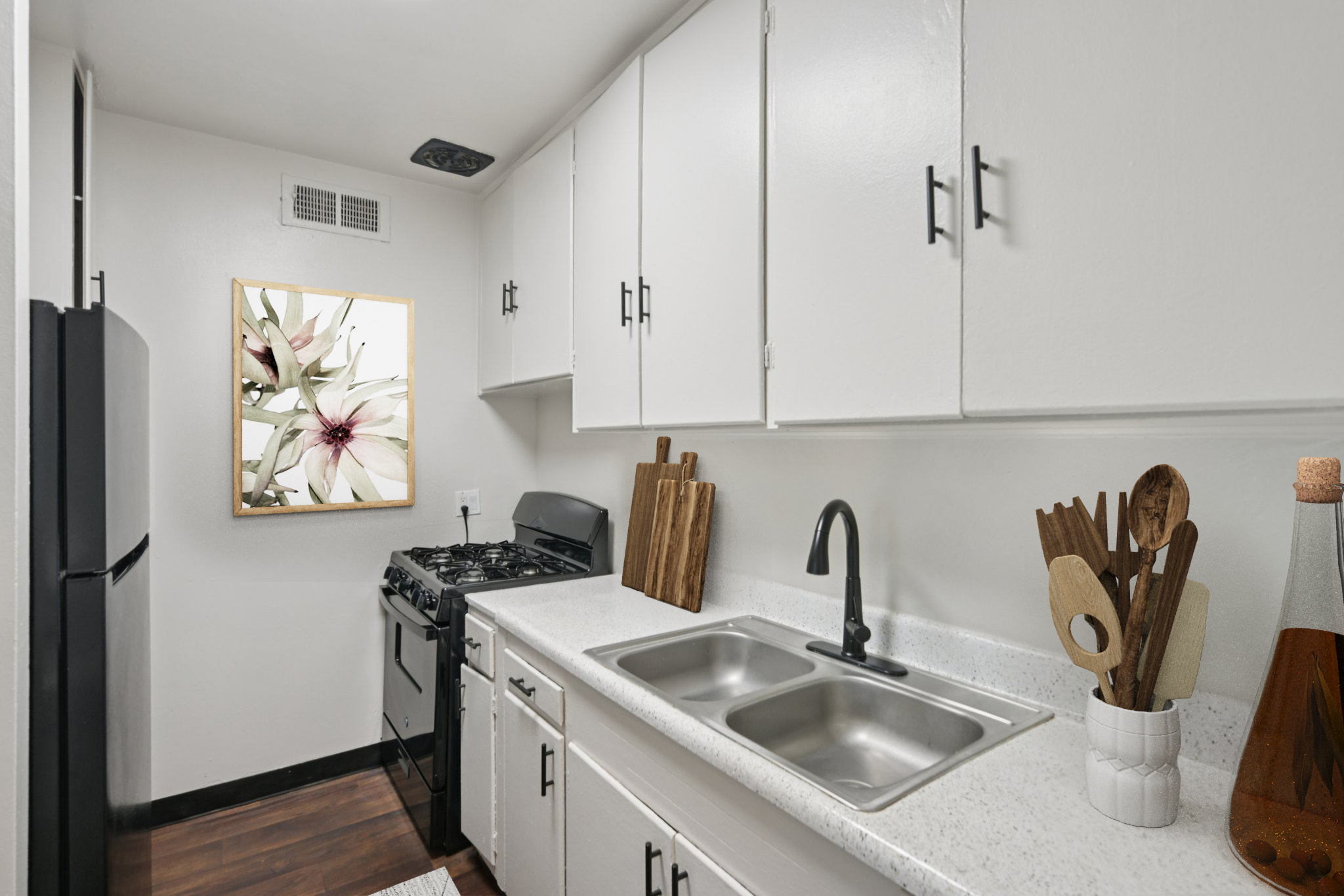 A modern kitchen featuring white cabinets, a black stove, a double sink, and wooden utensils. There is a framed image of flowers on the wall, and a stylish bottle on the counter, creating a clean and inviting atmosphere.