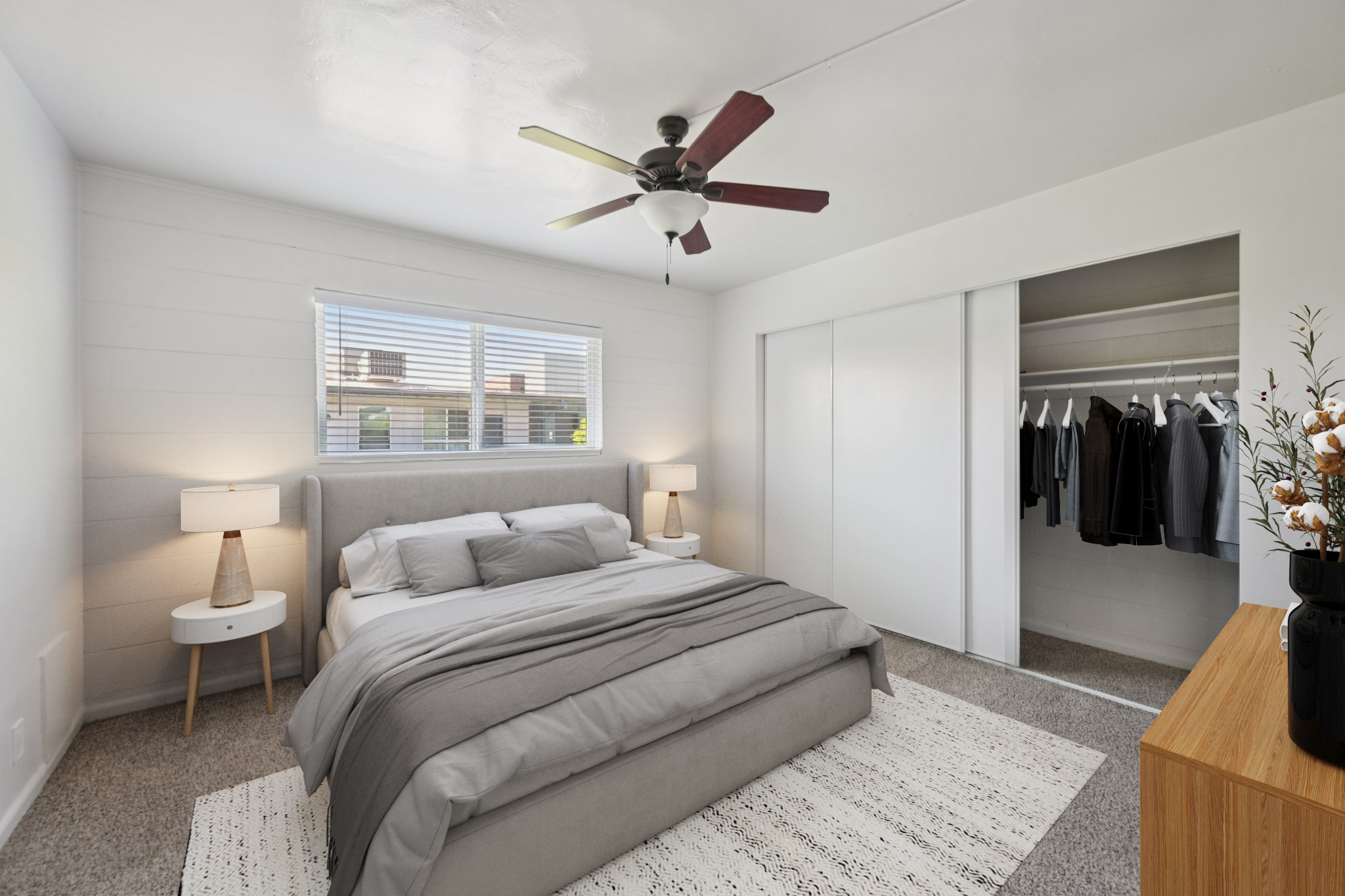 A modern bedroom featuring a queen-size bed with gray bedding, a nightstand with a lamp, and a ceiling fan. There is a large window with blinds allowing natural light, and a closet with hanging clothes. A decorative plant sits on a wooden dresser, enhancing the contemporary aesthetic.