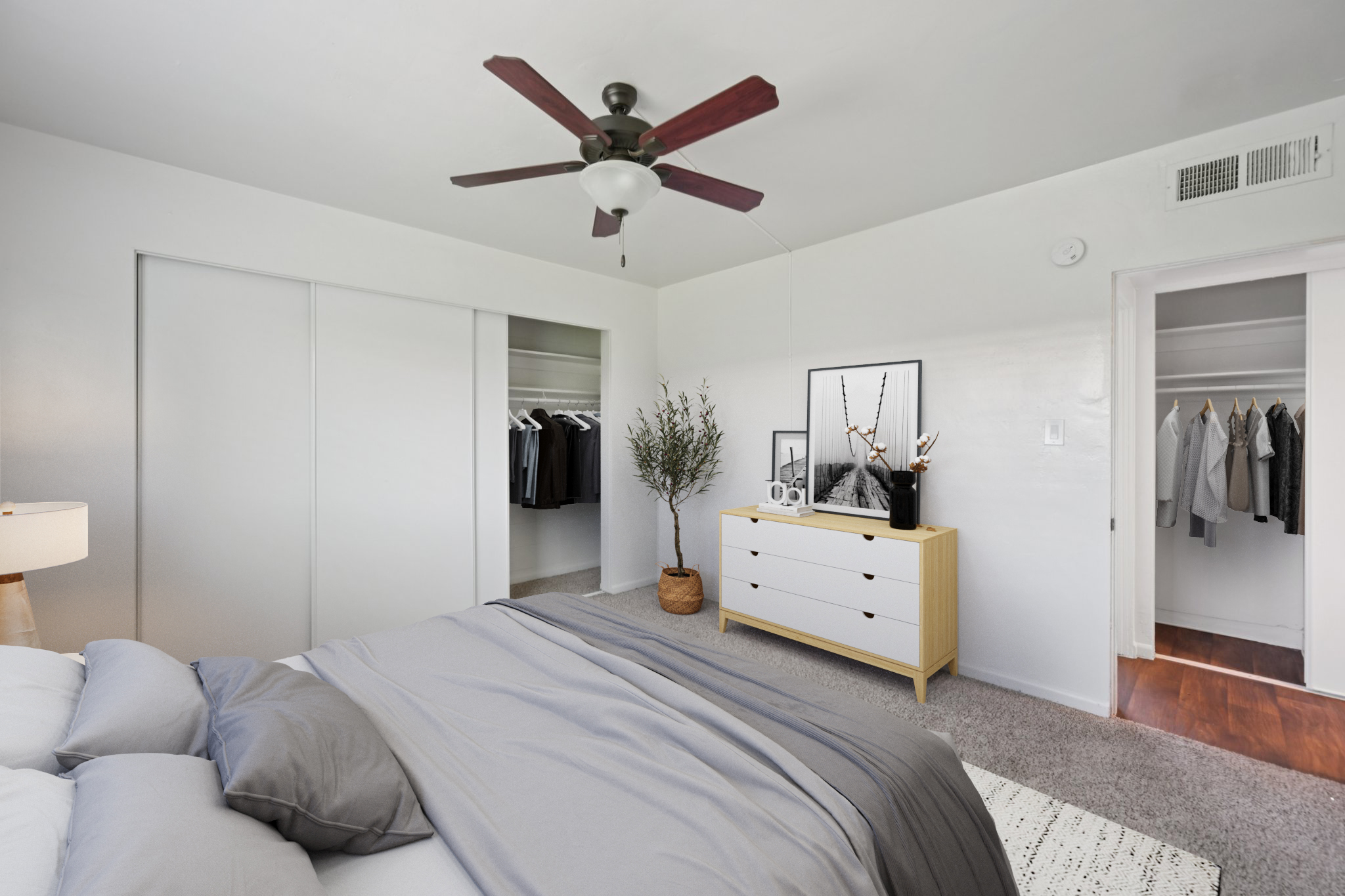 A well-decorated bedroom featuring a queen-sized bed with gray bedding, a white dresser with decorative items, and a ceiling fan. To the side, there's a closet with hanging clothes. The walls are light-colored, and there’s a warm ambient light from a lamp. The carpet adds a cozy touch to the space.