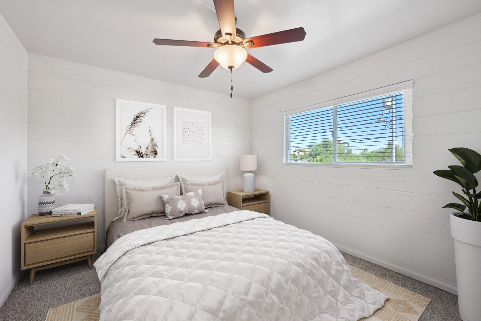 Cozy bedroom featuring a queen-sized bed with a light gray comforter, two bedside tables with lamps, and a decorative plant. Natural light streams through a window with blinds, complemented by a ceiling fan. Simple wall art adds a touch of style to the white walls.