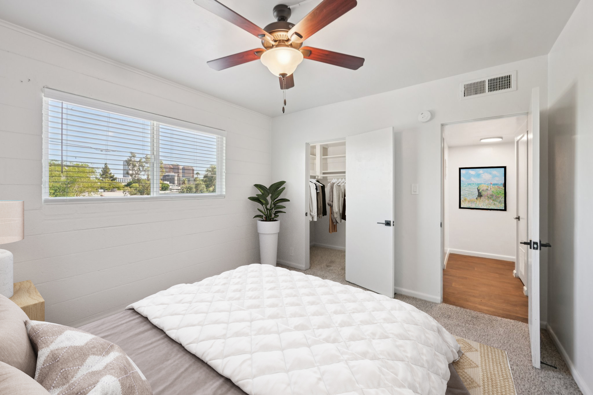 A cozy bedroom featuring a comfortable bed with a fluffy white comforter, a ceiling fan, and a large window allowing natural light. A potted plant sits beside the bed, and a closet is visible through an open door. The room has neutral-colored walls and soft carpeting, creating a warm and inviting atmosphere.