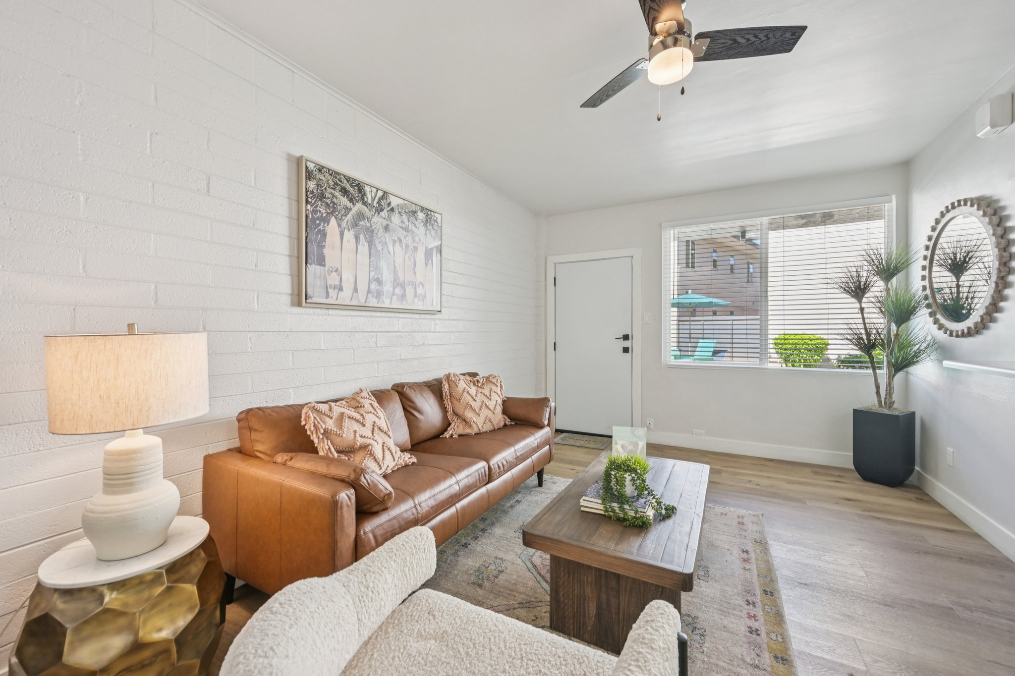 A modern living room featuring a brown leather sofa with decorative pillows, a wooden coffee table, and a stylish lamp. A large window lets in natural light, displaying plants outside. A round mirror hangs on the wall, and a ceiling fan is visible above. The decor is minimalist and inviting.